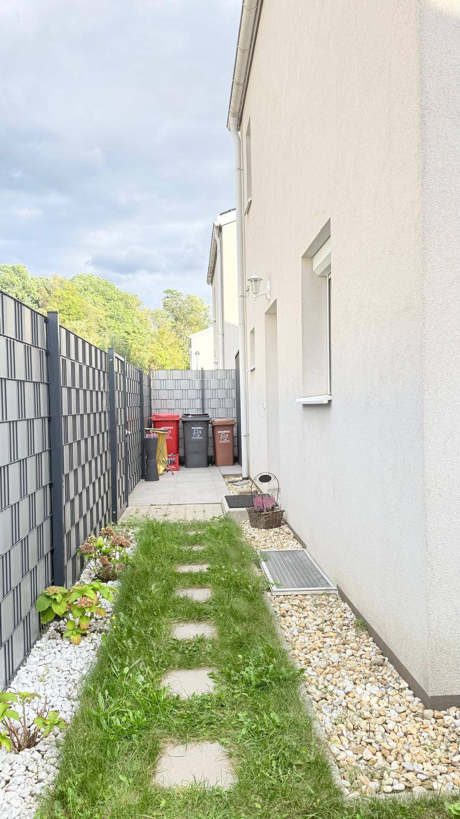 Seitlicher Zugangsweg am Haus mit Kiesstreifen, Trittsteinen und einem modernen Zaun.