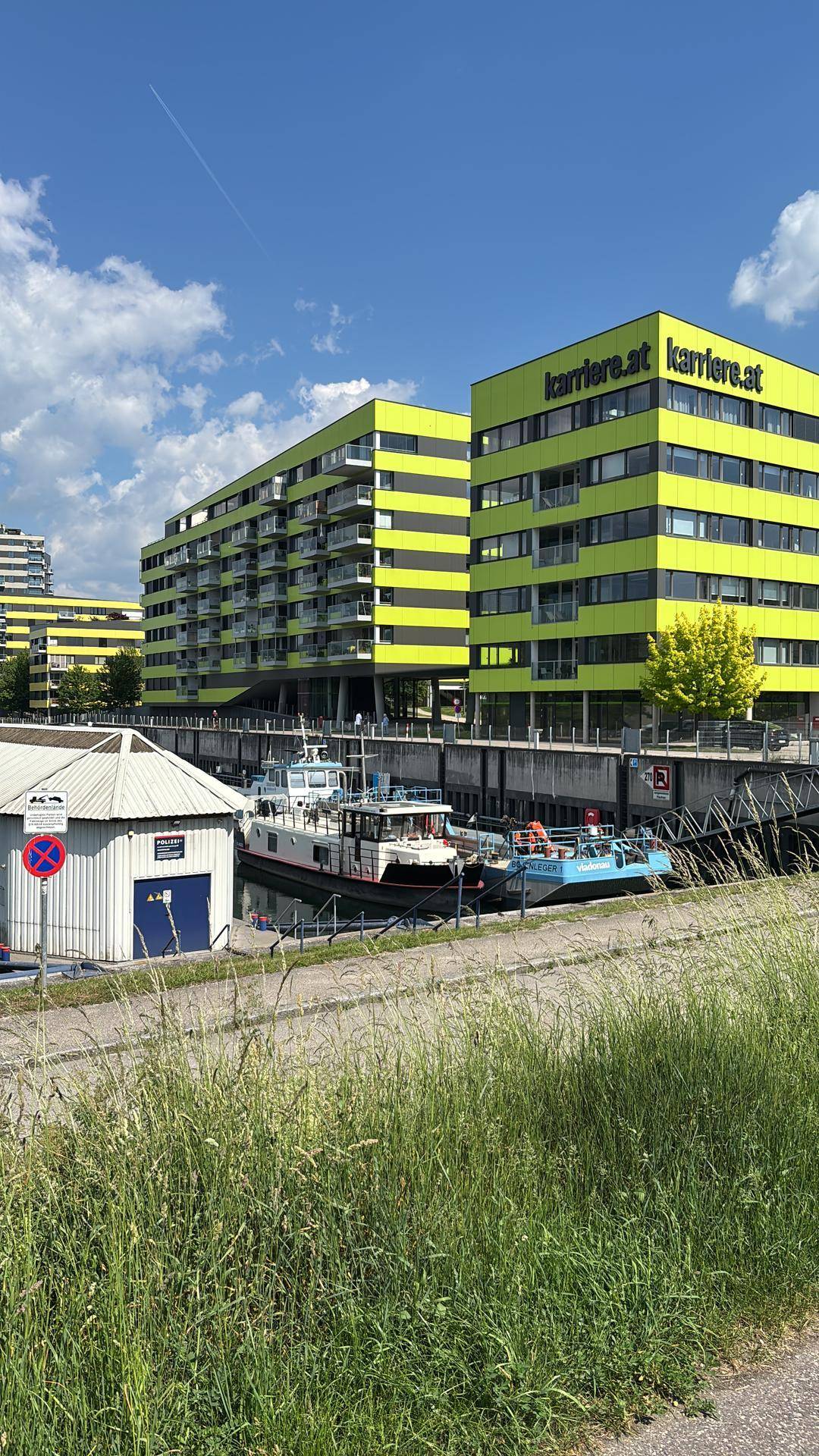 Moderne gelbe Gebäude mit Balkonen am Wasser, umgeben von grüner Natur unter einem klaren blauen Himmel.