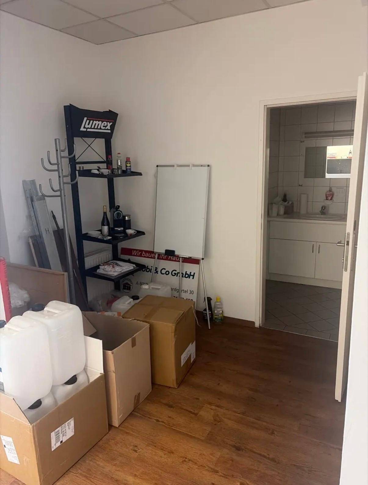 Ecke eines Raumes mit Regalen, Kartons und Whiteboard, mit Zugang zu einem gefliesten Badezimmer.
