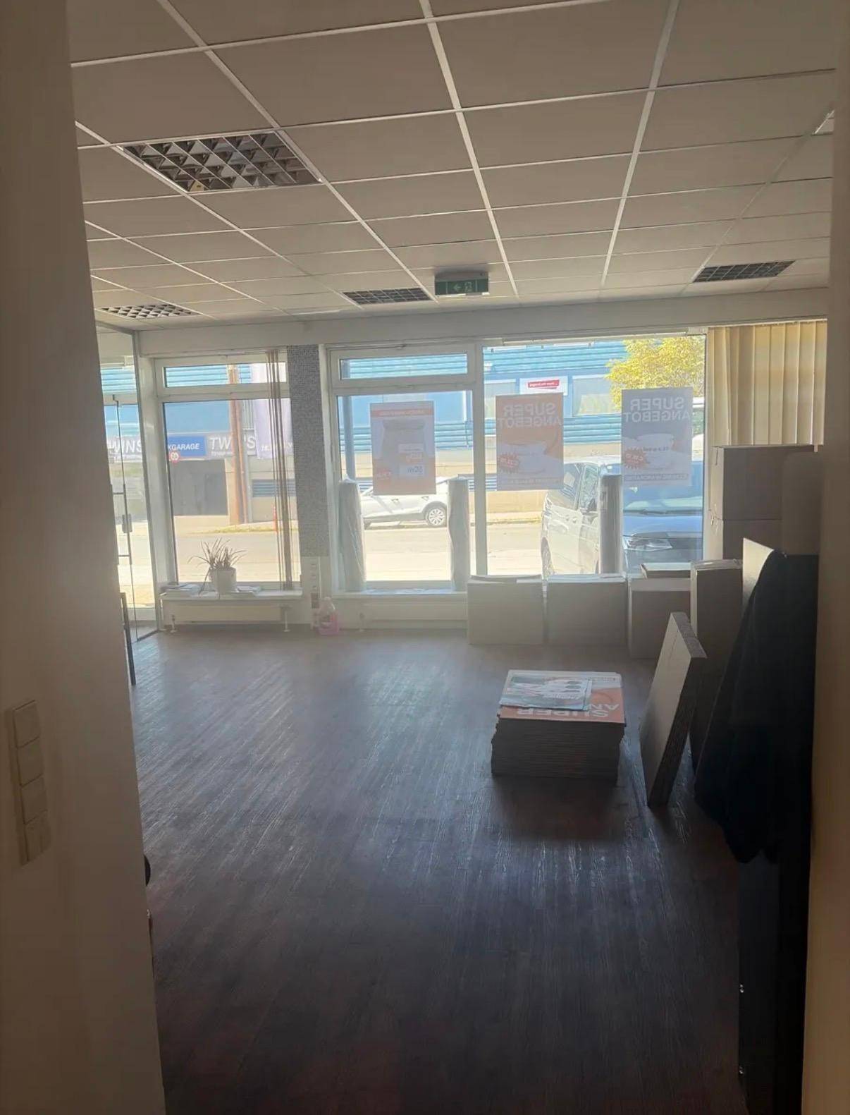 Heller, offener Raum mit großen Fenstern zur Straße, Laminatboden und abgehängter Decke, teilweise mit Kartons.