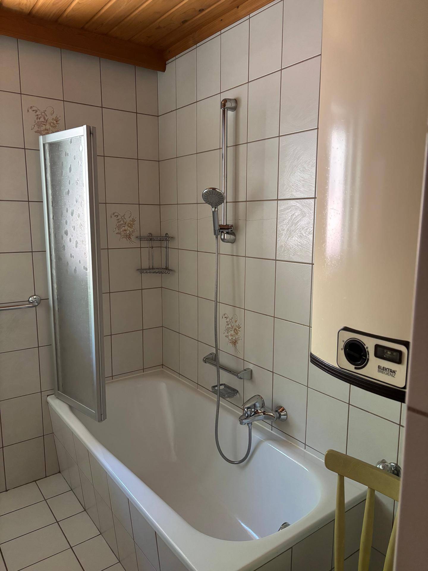 Badewanne mit Duscharmatur und Duschwand, umgeben von weißen Fliesen und einem Warmwasserboiler.