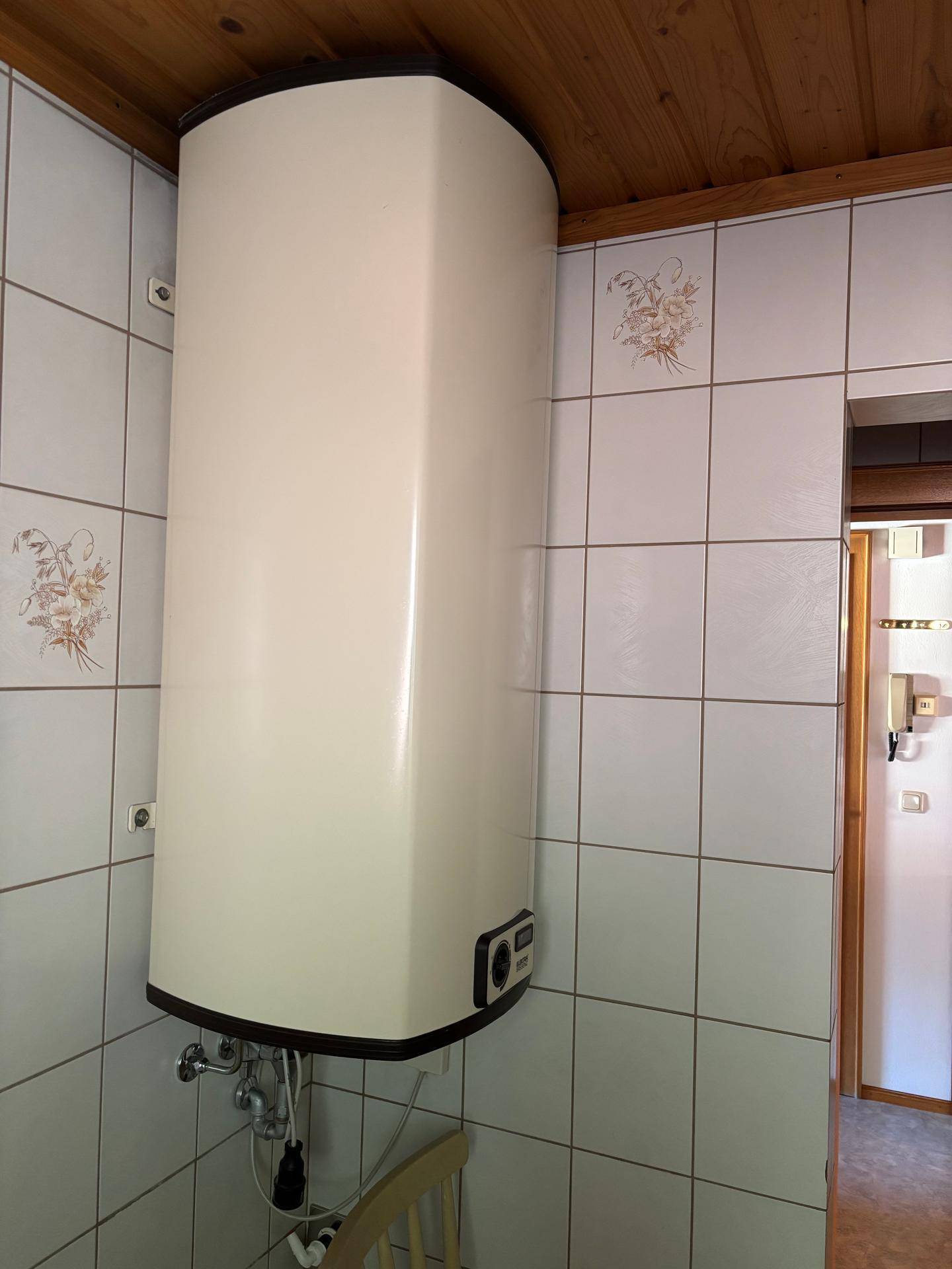 Wandmontierter Warmwasserboiler im Badezimmer, umgeben von weißen Fliesen und Holzdecke.
