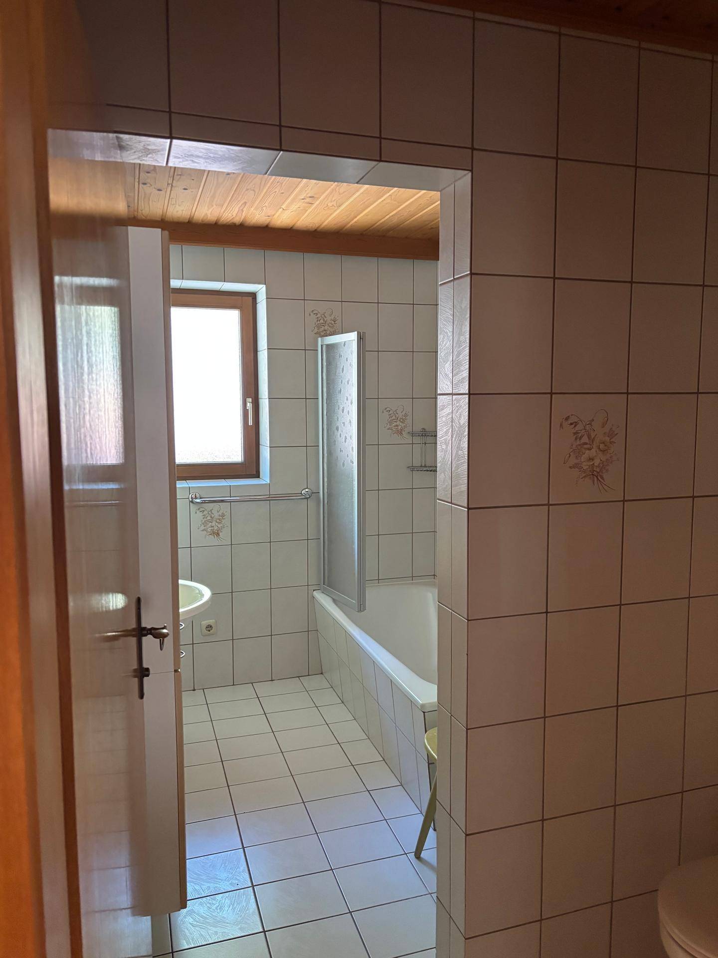 Badezimmer mit weißen Fliesen, Badewanne mit Duschwand und einem Fenster für Tageslicht.