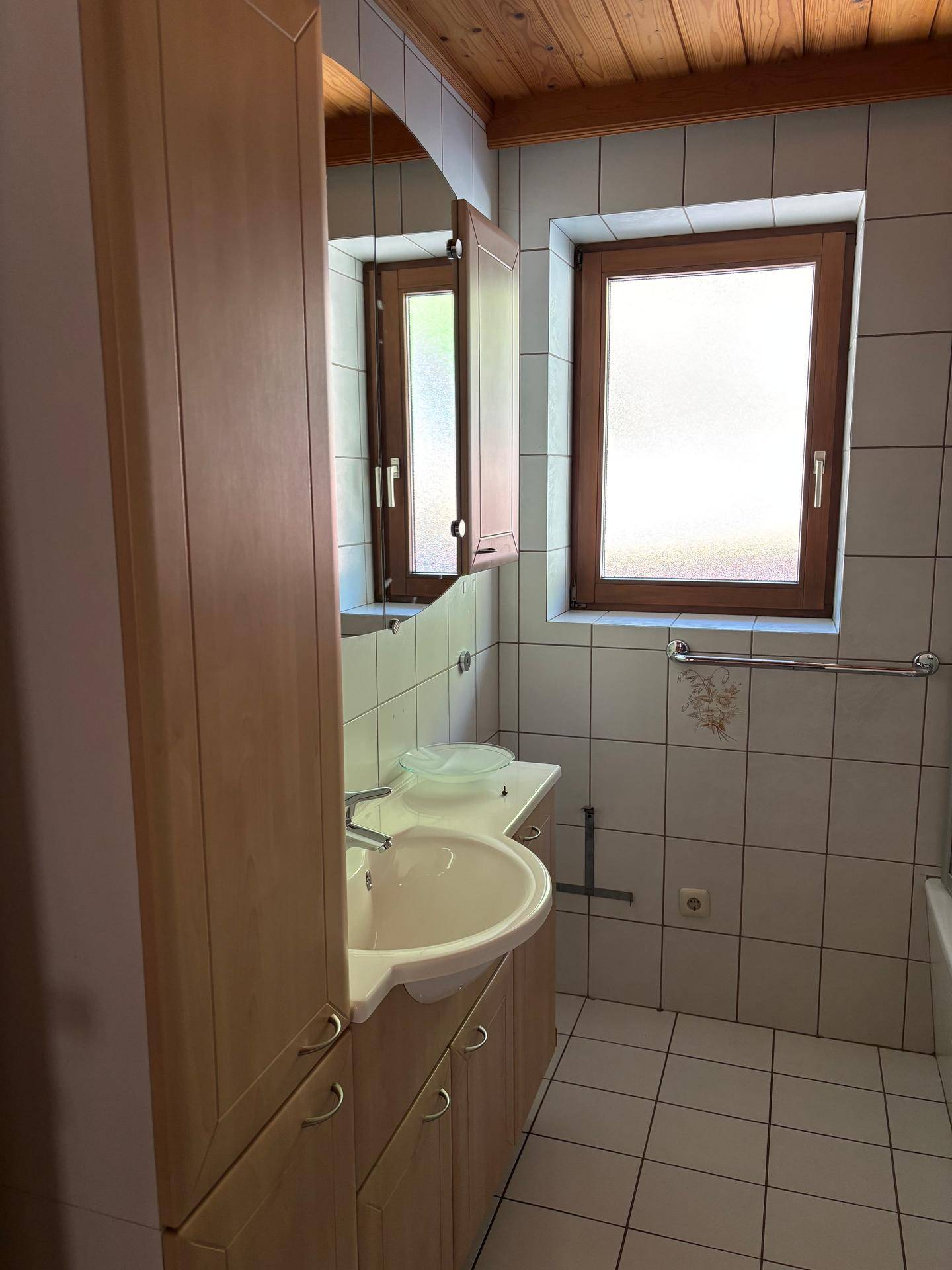 Badezimmer mit Waschtisch, Spiegelschrank, weißen Fliesen und einem Fenster für natürliche Belüftung.