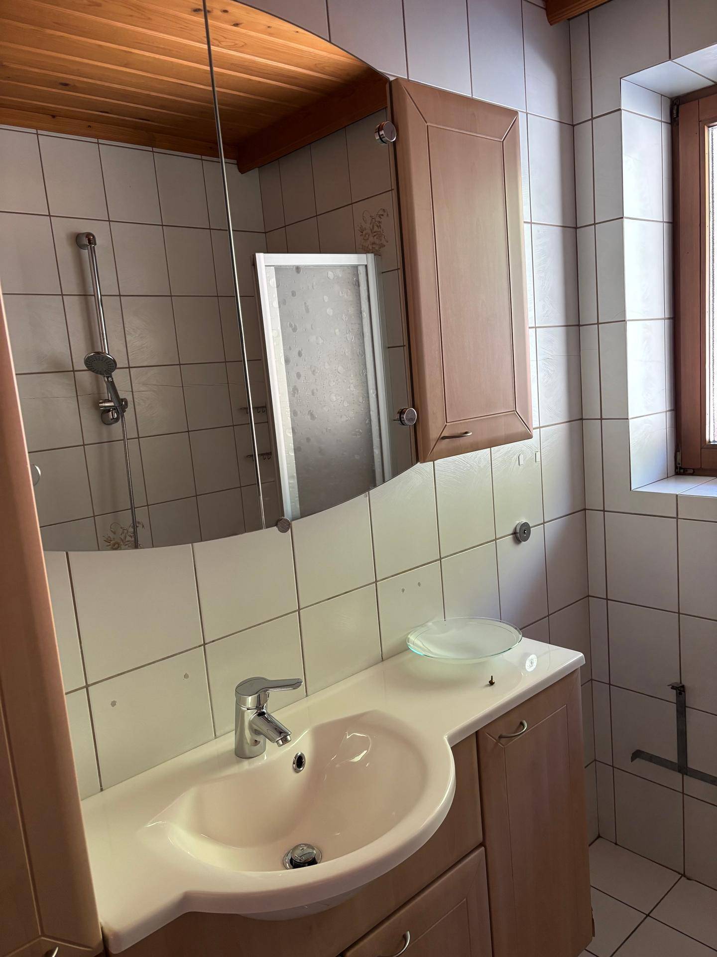 Badezimmerwaschtisch mit großem Spiegel, weißen Fliesen und einem Fenster für helles Tageslicht.