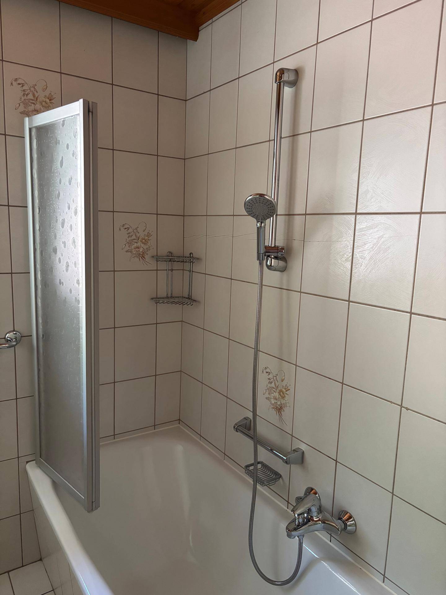Badewanne mit Duscharmatur und Duschwand, umgeben von weißen Fliesen und traditioneller Holzdecke.