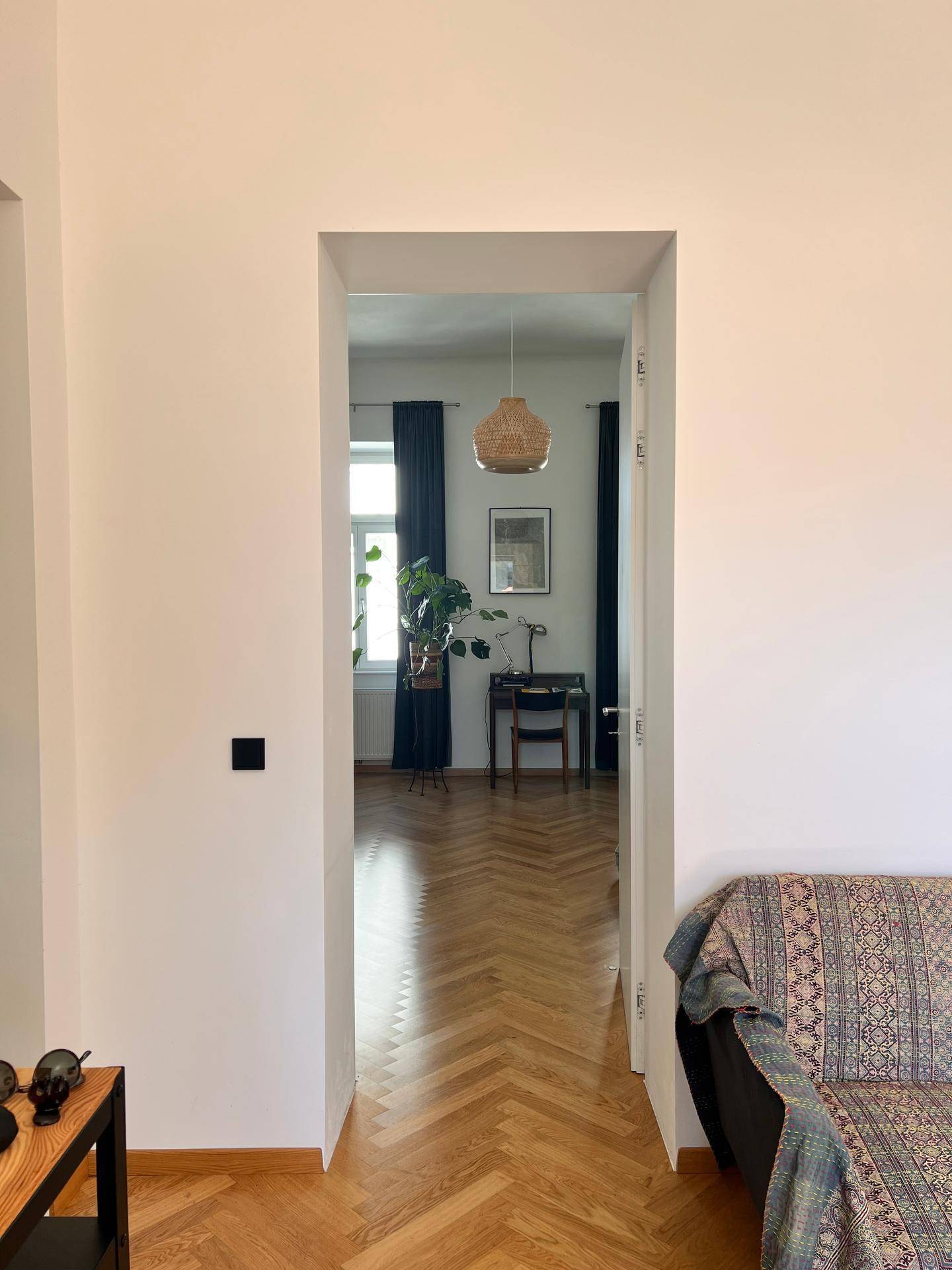 Blick durch eine Türöffnung in ein helles Zimmer mit Fischgrätparkett und großen Fenstern.