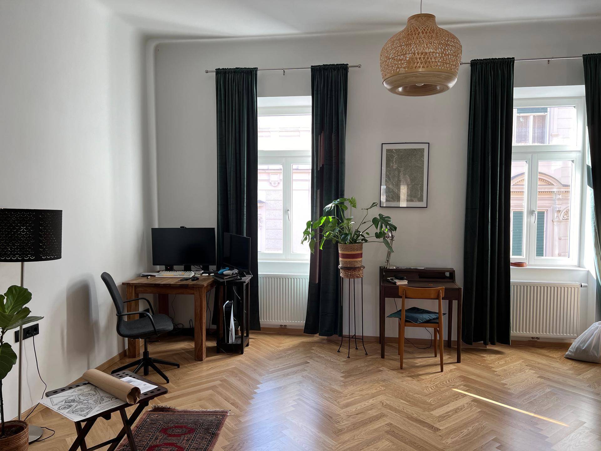 Helles Wohnzimmer mit Fischgrätparkett, zwei großen Fenstern und einem Arbeitsbereich.