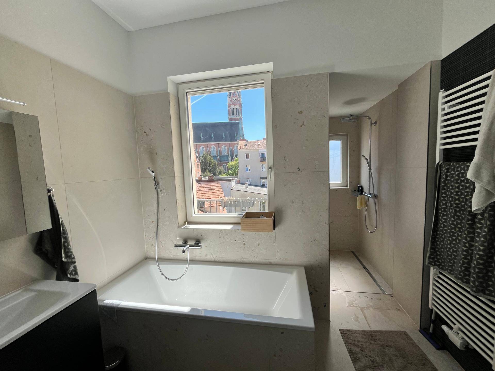 Helles Badezimmer mit Badewanne, Dusche und Fenster mit Blick auf einen Kirchturm.