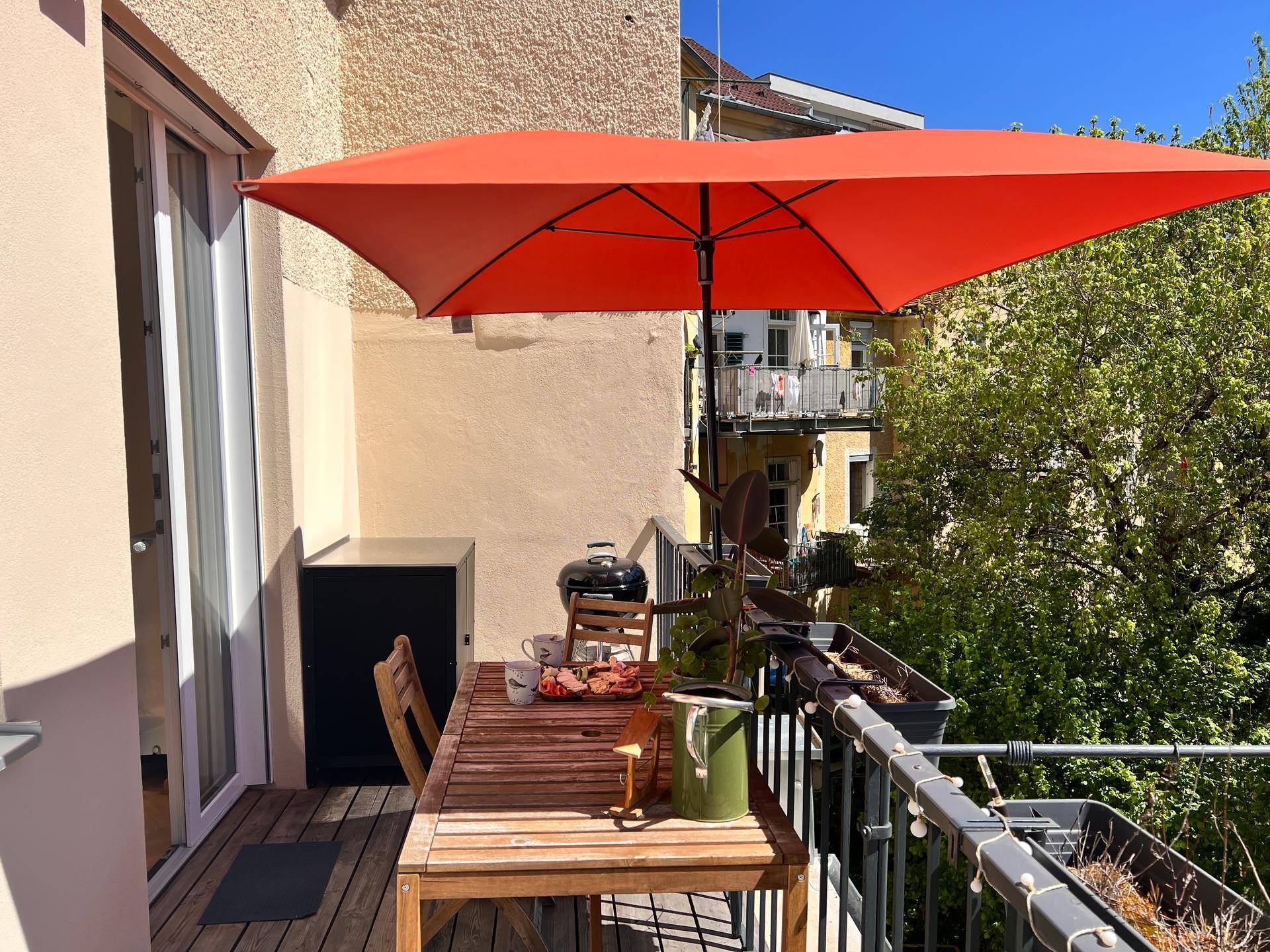 Gemütlicher Balkon mit orangefarbenem Sonnenschirm, Holztisch und Grill für entspannte Stunden.