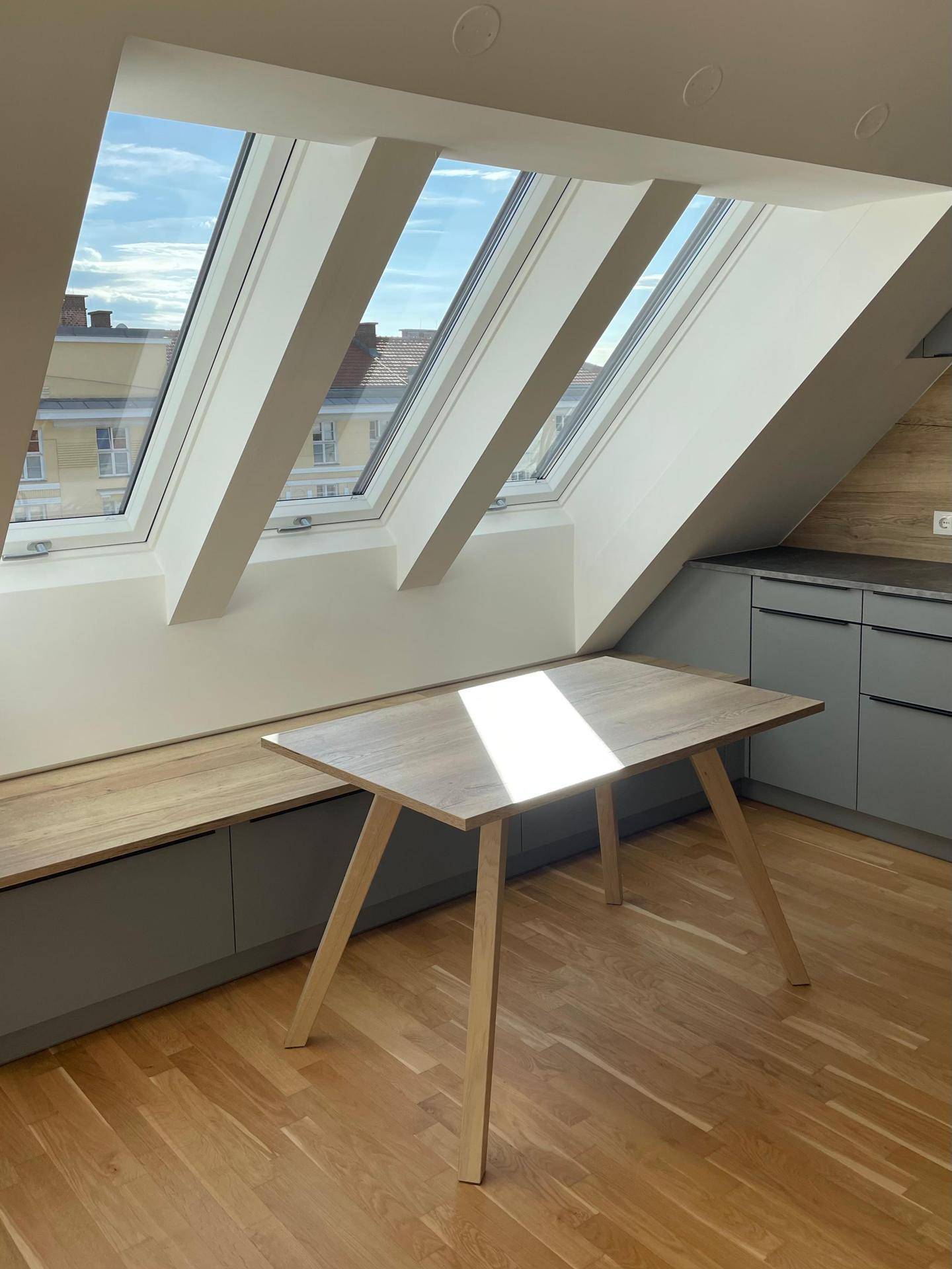Heller Küchenbereich im Dachgeschoss mit großen Dachfenstern, Holzboden und modernen grauen Schränken.