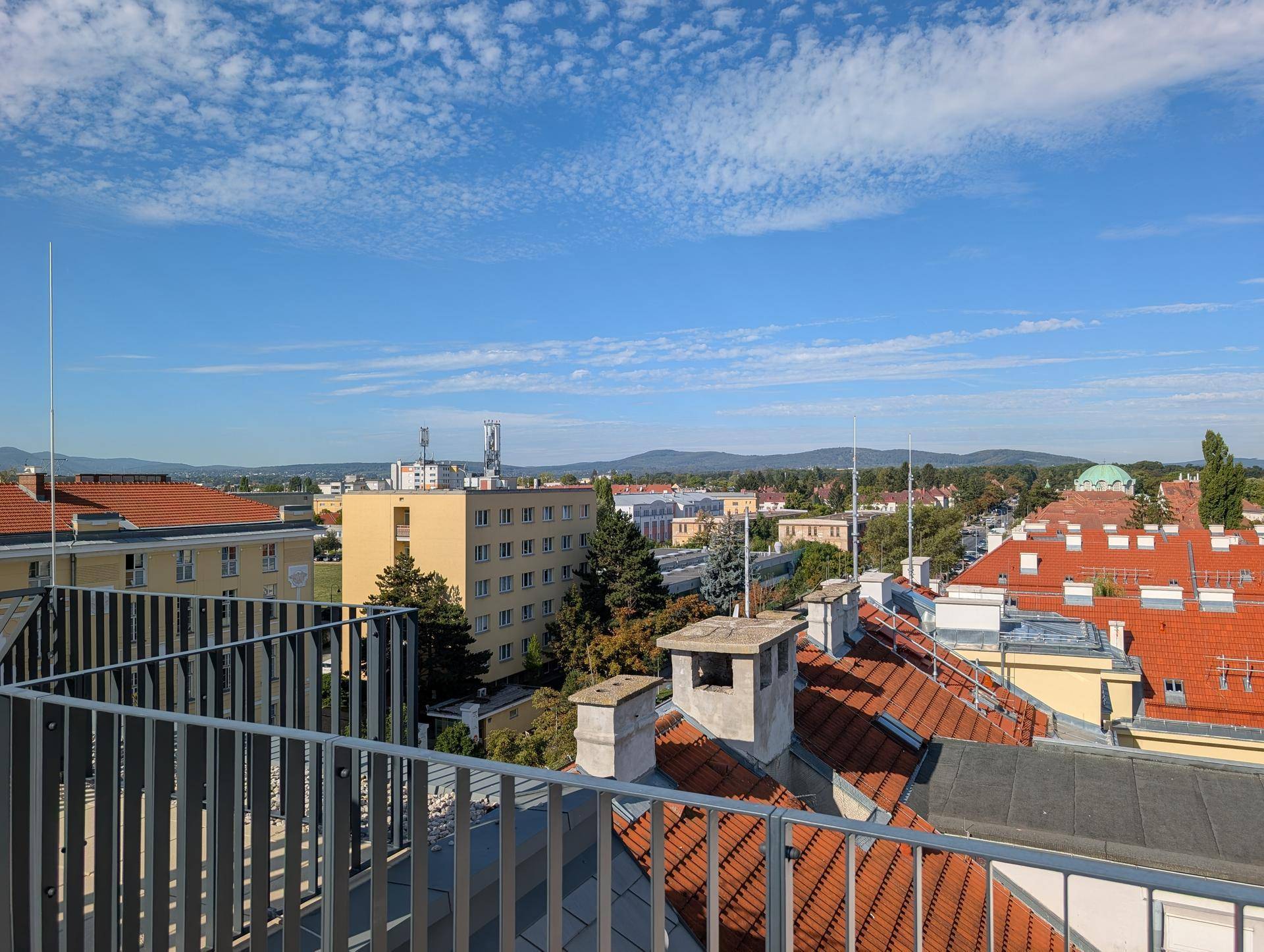 Weitläufiger Ausblick über die Dächer der Stadt bis zu den grünen Hügeln am Horizont unter blauem Himmel.