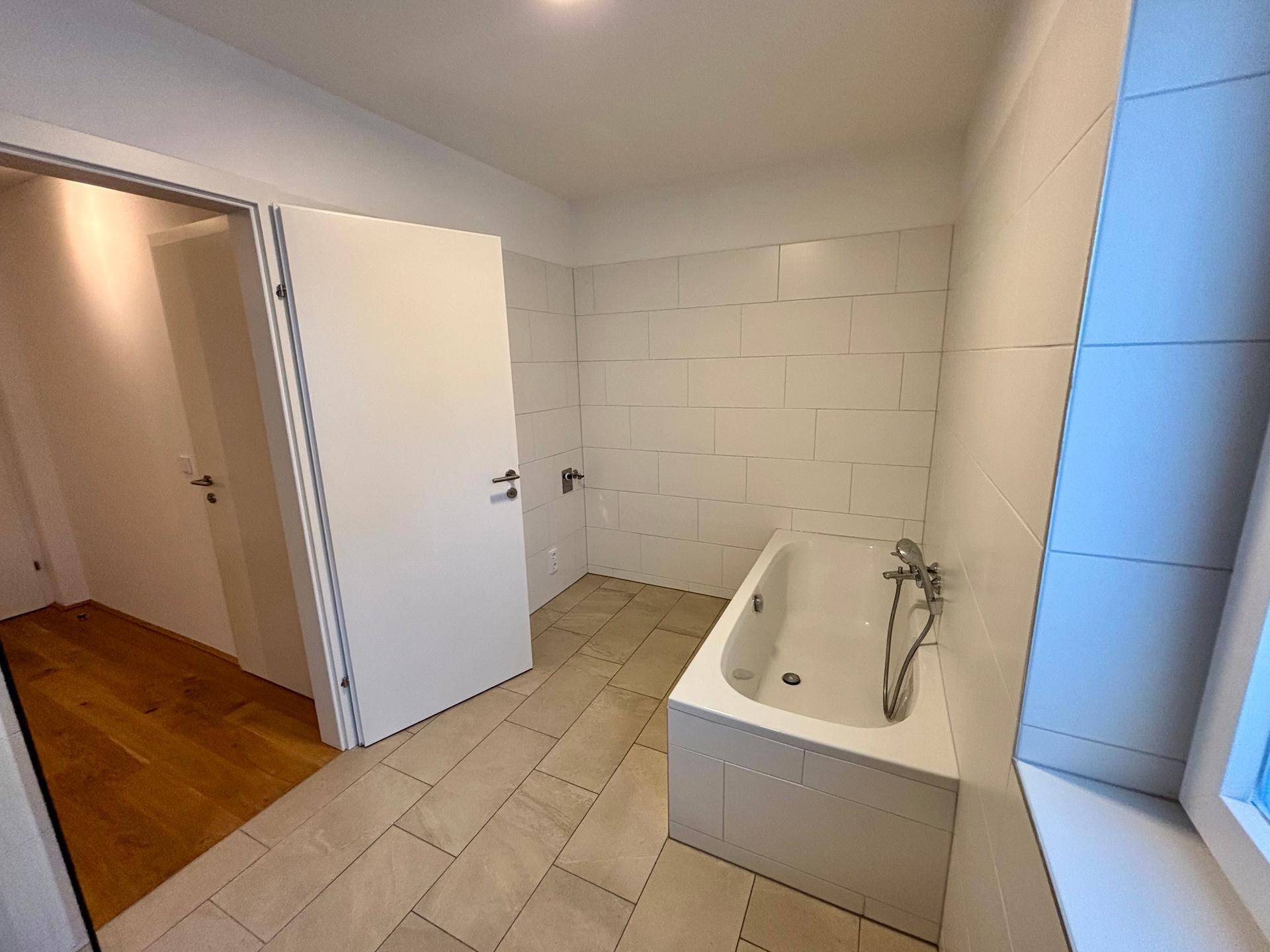 Badezimmer mit Badewanne, weißen Fliesen und Zugang zu einem weiteren Raum.