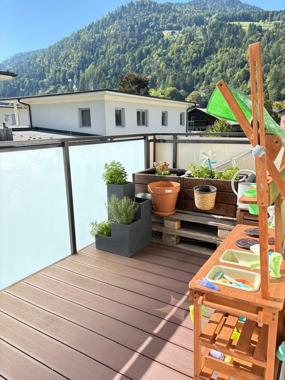 Sonnige Terrasse mit Holzboden, Kräuterbeeten und Spielbereich für Kinder mit Bergblick.