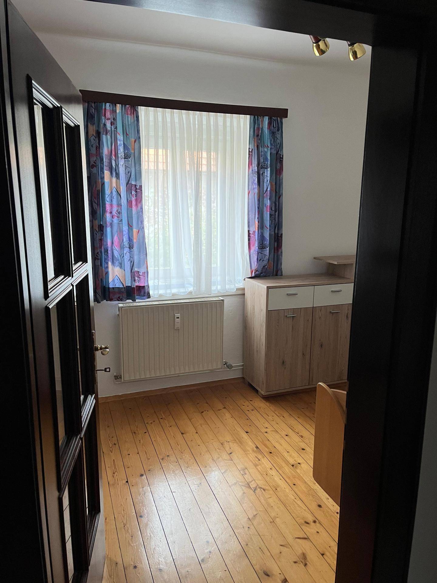 Helles Zimmer mit Parkettboden, Fenster und bunten Vorhängen.