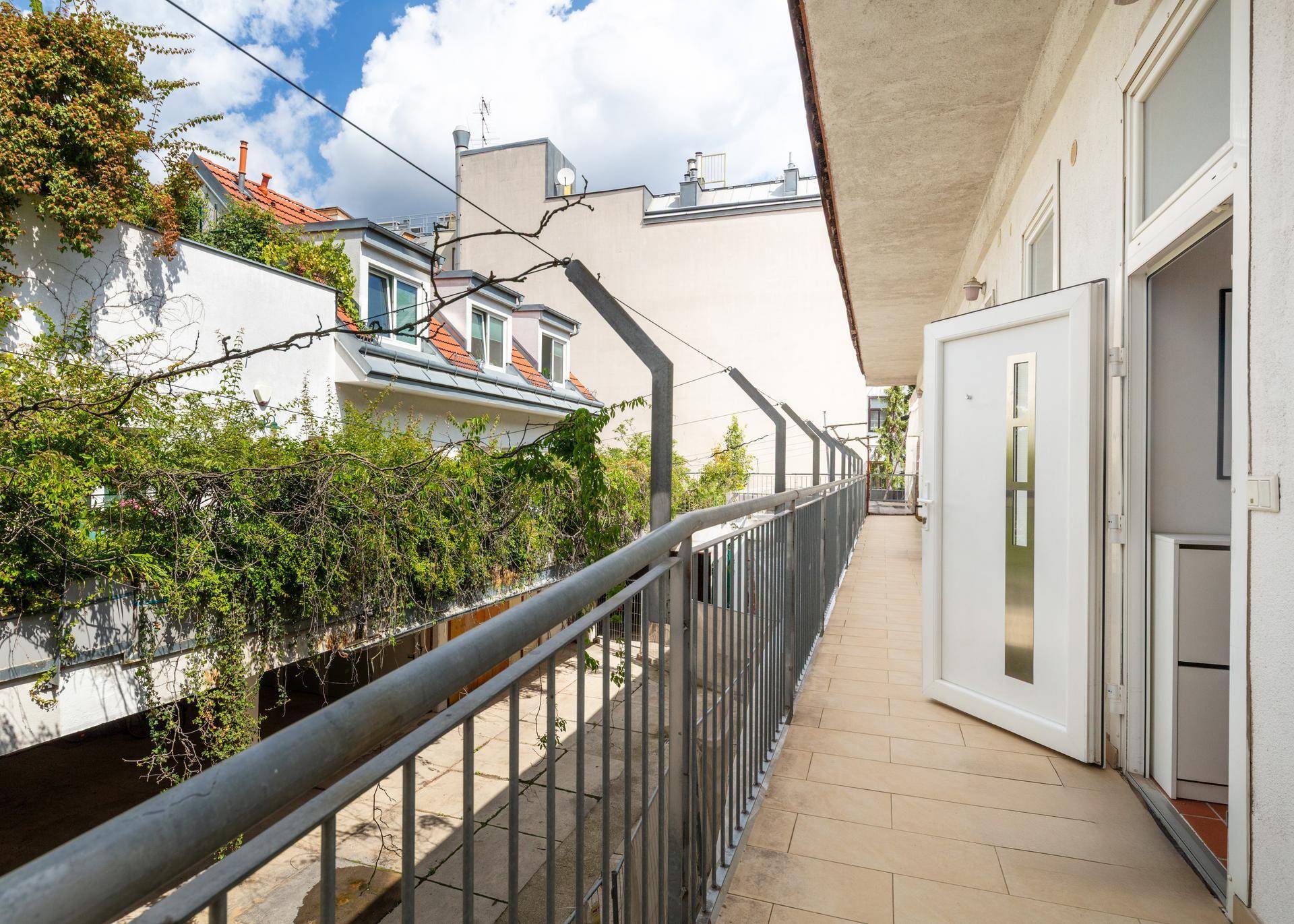 Langer Balkon mit Metallgeländer und Blick auf begrünte Dächer und Nachbargebäude.