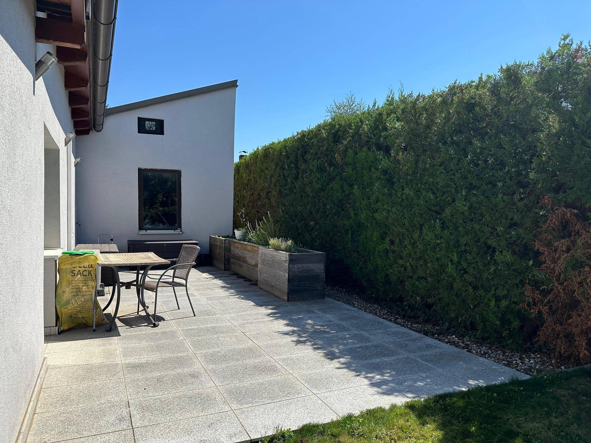 Sonniger Terrassenbereich mit Steinplatten, Gartenmöbeln und hoher Hecke für Privatsphäre.