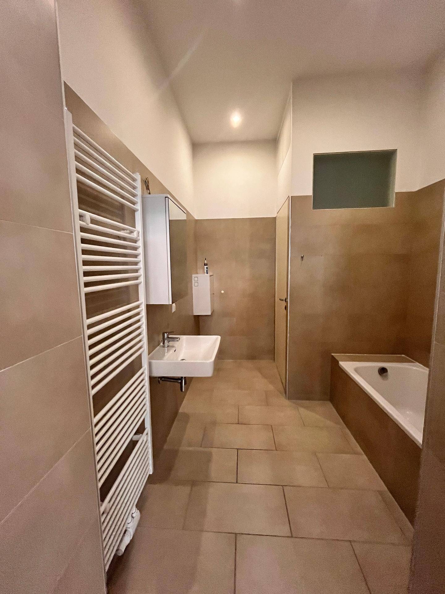 Modernes Badezimmer mit beigefarbenen Fliesen, Badewanne, Einzelwaschbecken und Handtuchheizkörper.