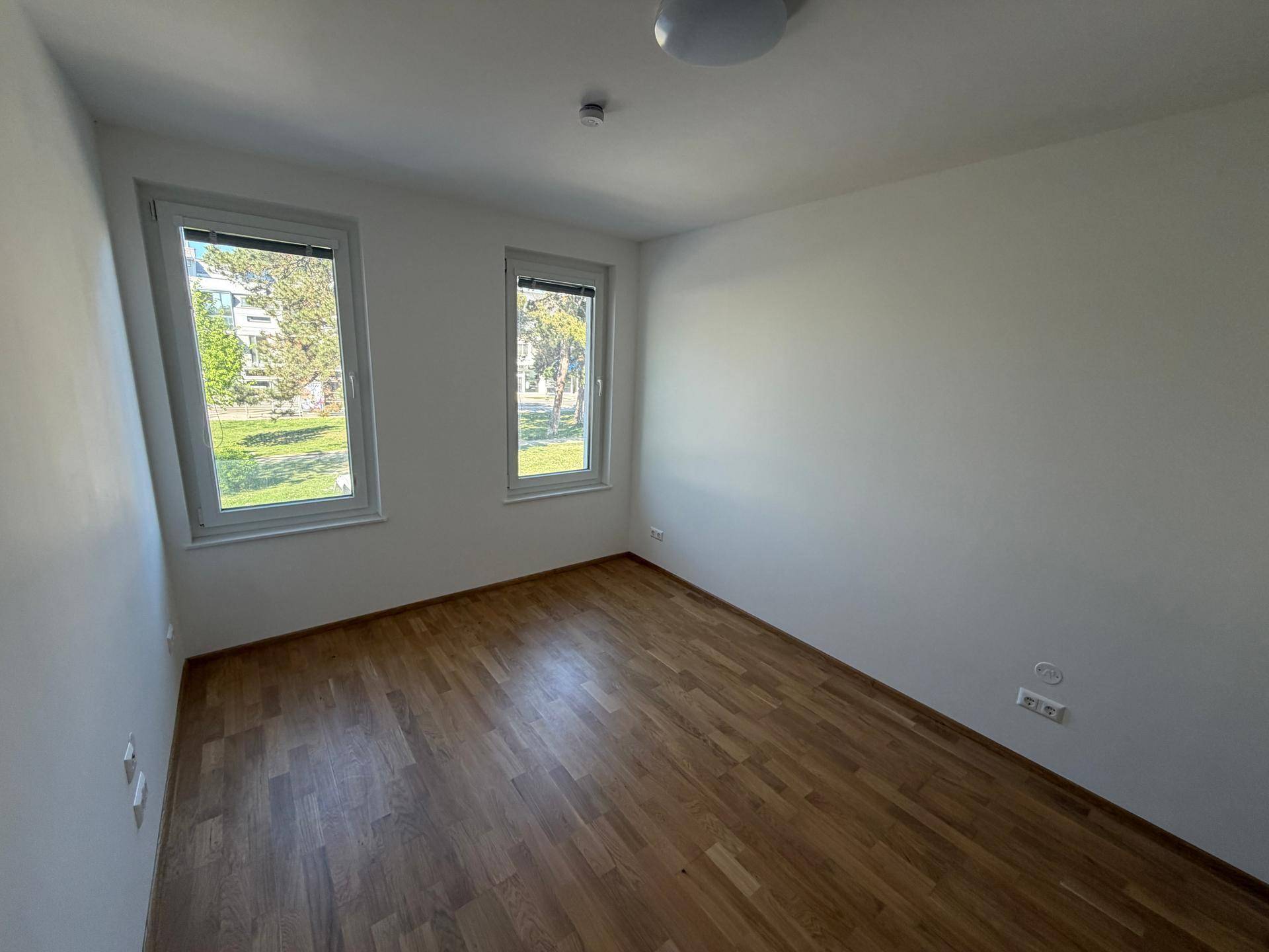 Leeres Zimmer mit Holzboden, weißen Wänden und zwei Fenstern mit Blick ins Grüne.