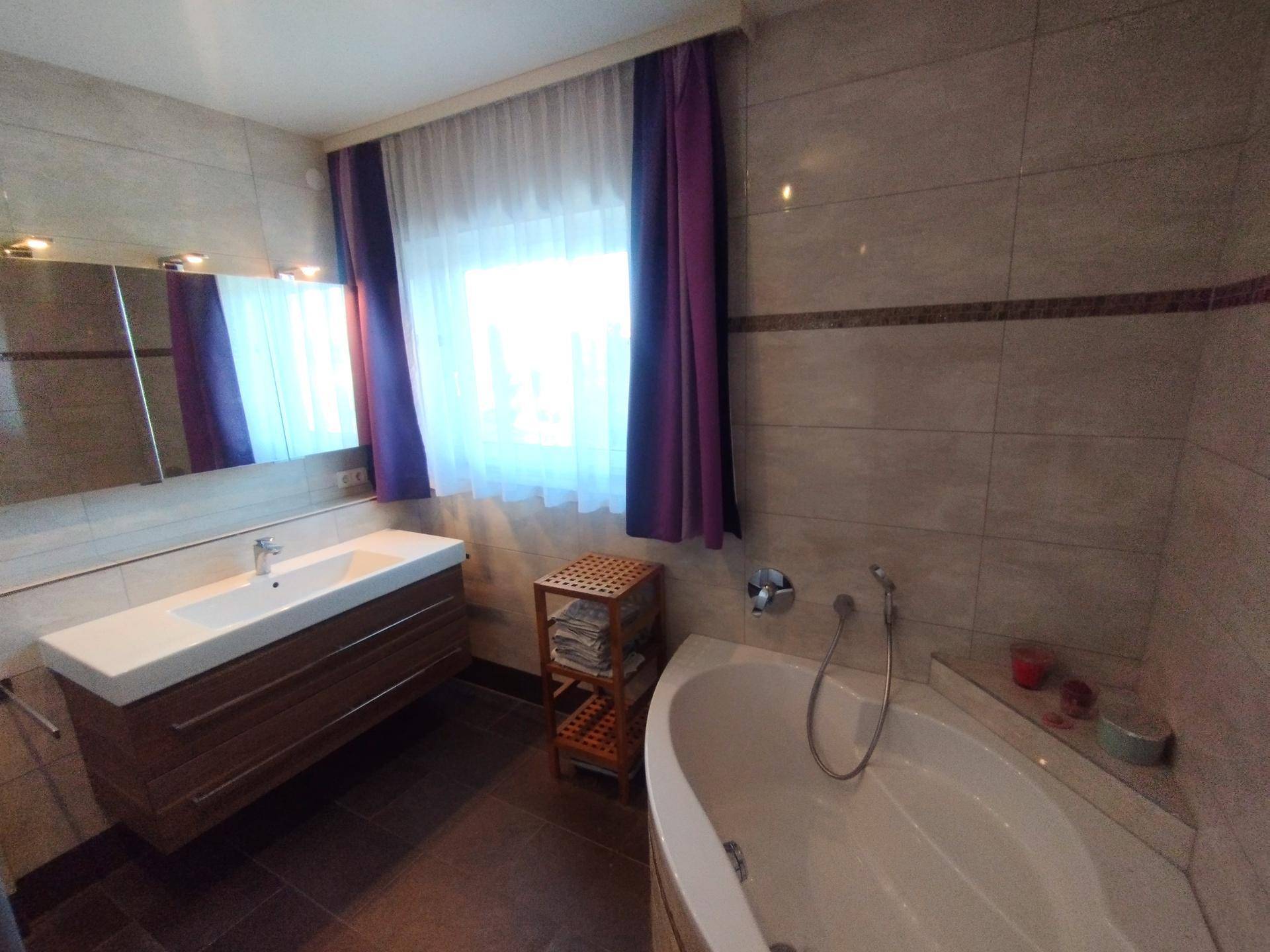 Gefliestes Badezimmer mit Eckbadewanne, modernem Waschtisch, Spiegelschrank und Fenster.