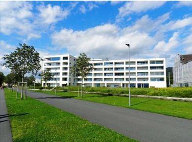 Modernes weißes Apartmentgebäude mit vielen Balkonen und großen Fenstern unter blauem Himmel.