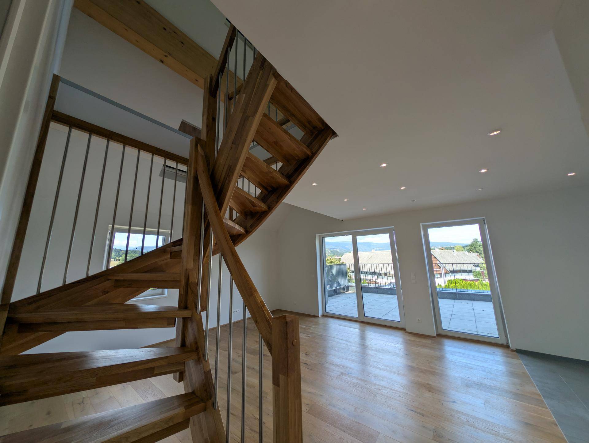 Elegante Holztreppe führt zum hellen Wohnbereich mit großen Fenstern und Blick ins Grüne.
