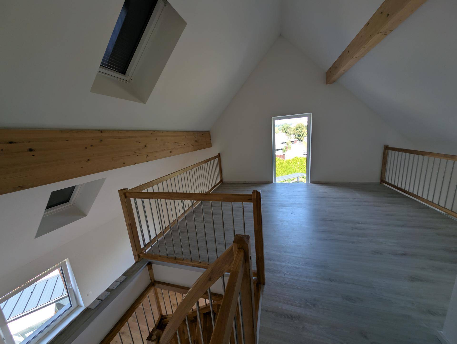 Helle Galerie im Dachgeschoss mit sichtbaren Holzbalken, Dachfenstern und Zugang nach draußen.