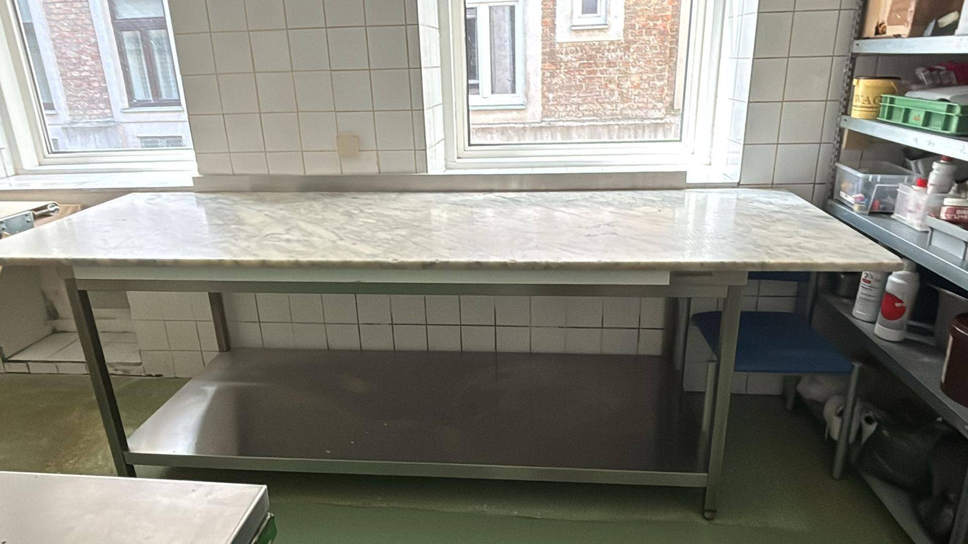 Großer Arbeitstisch mit Marmorplatte und Edelstahlgestell neben einem Fenster in einer gewerblichen Küche.