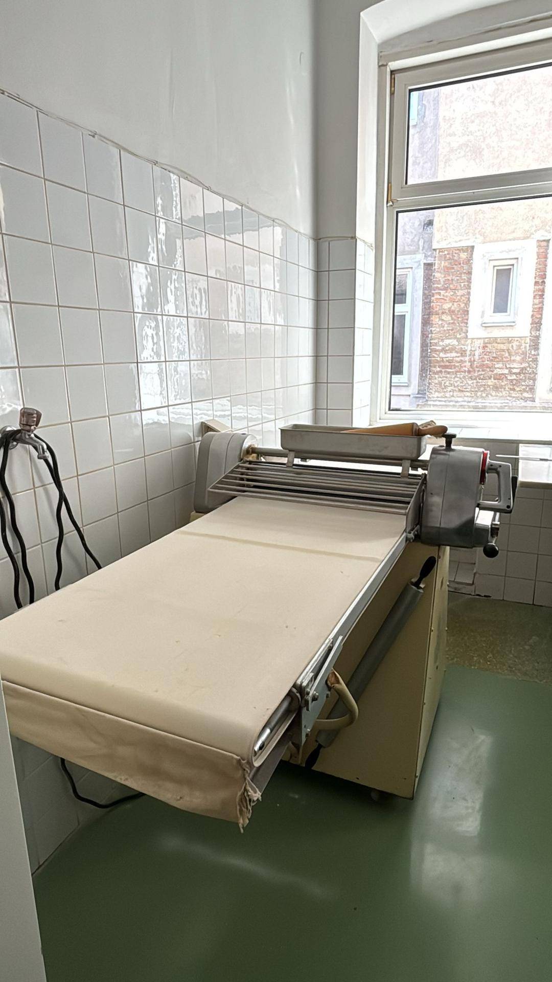 Eine Teigausrollmaschine steht neben einem Fenster in einer professionellen Küche mit weißen Fliesenwänden und grünem Boden.