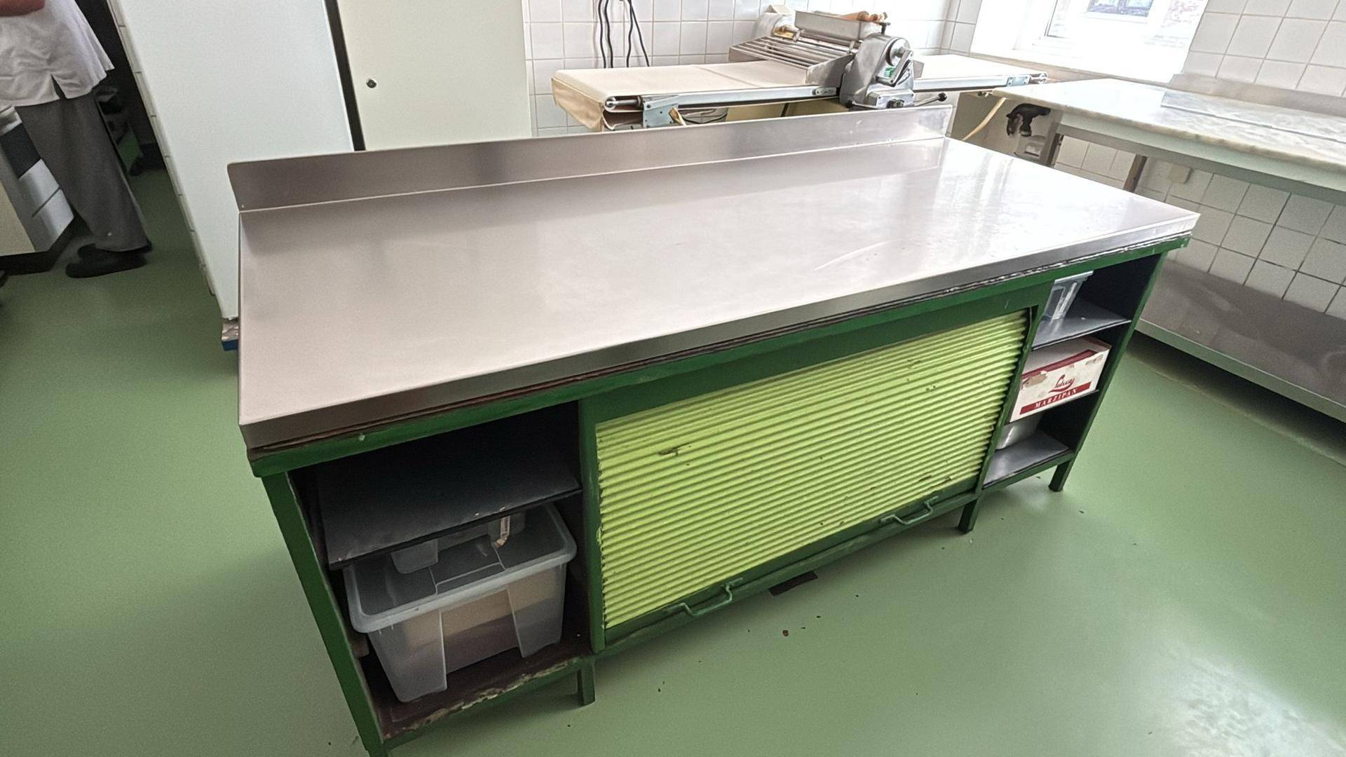 Arbeitsinsel mit Edelstahlplatte und grünem Rollladenschrank in einer gewerblichen Küche mit Teigausrollmaschine im Hintergrund.