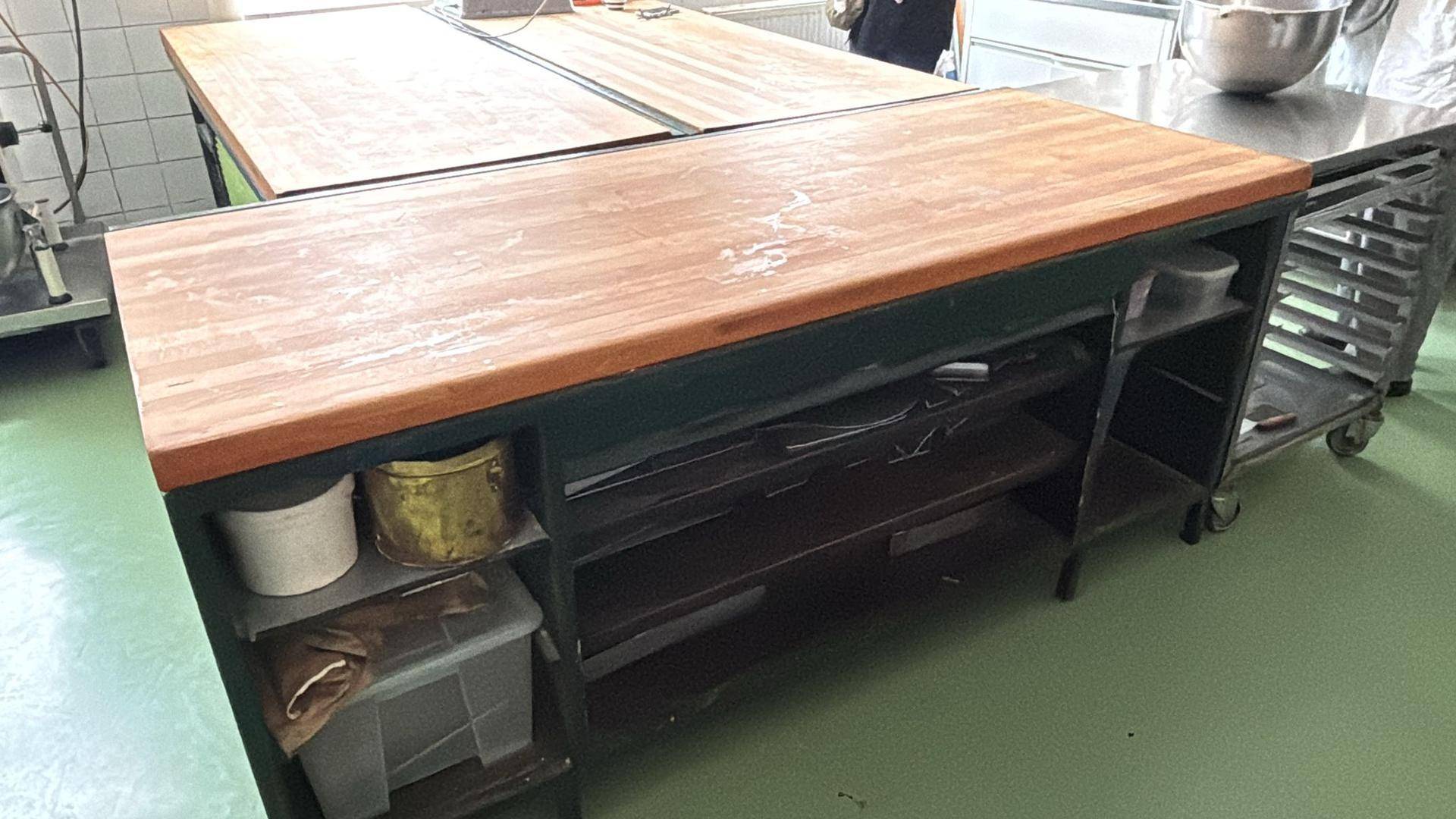 Eine große Arbeitsinsel mit Holzplatte und Regalen in einer gewerblichen Küche mit grünem Boden.