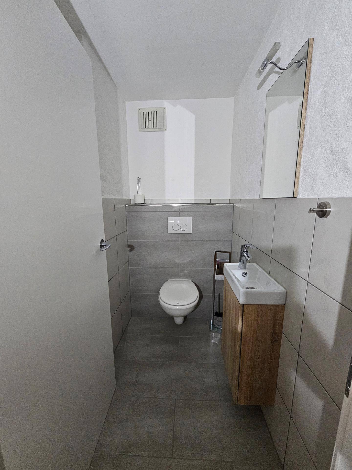 Kleines Gäste-WC mit Wand-WC, Waschbecken und grauen Fliesen.