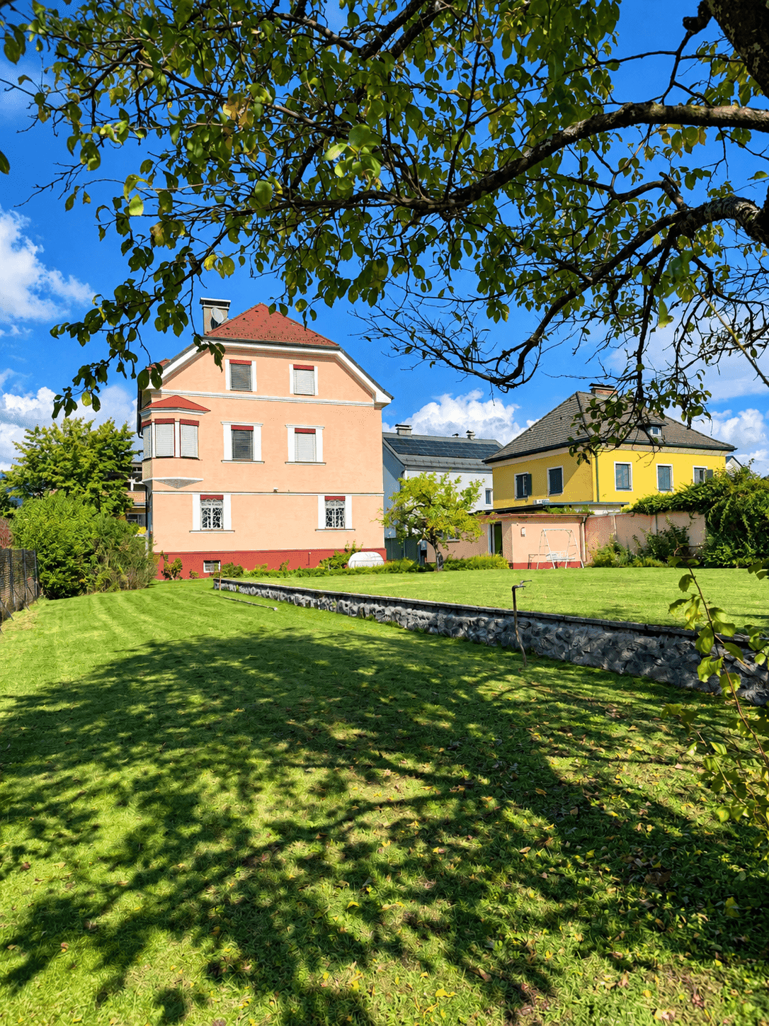 Großzügiger Gartenbereich mit altem Baumbestand und gepflegtem Rasen vor dem Haus unter blauem Himmel.