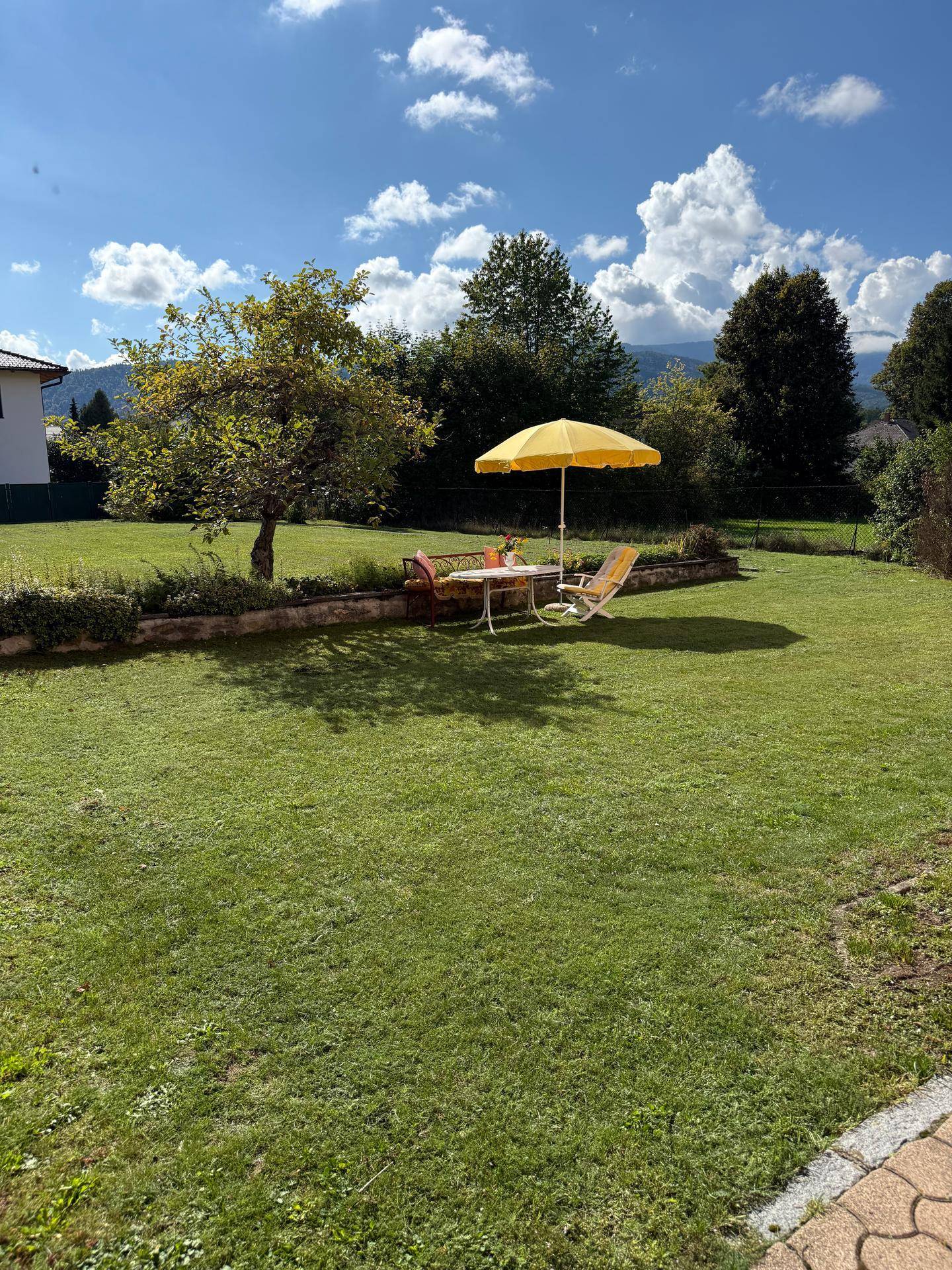 Weitläufiger Garten mit Rasenfläche, einem Baum und einer möblierten Sitzecke, umgeben von grüner Natur.