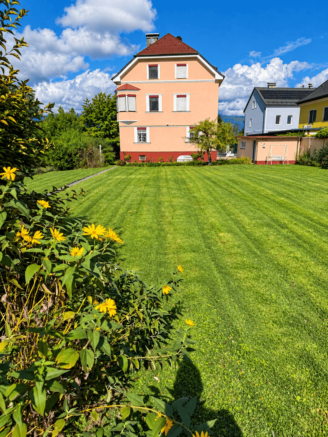 Ansicht des Hauses mit weitem Rasen und gelben Blumen im Vordergrund unter blauem Himmel.