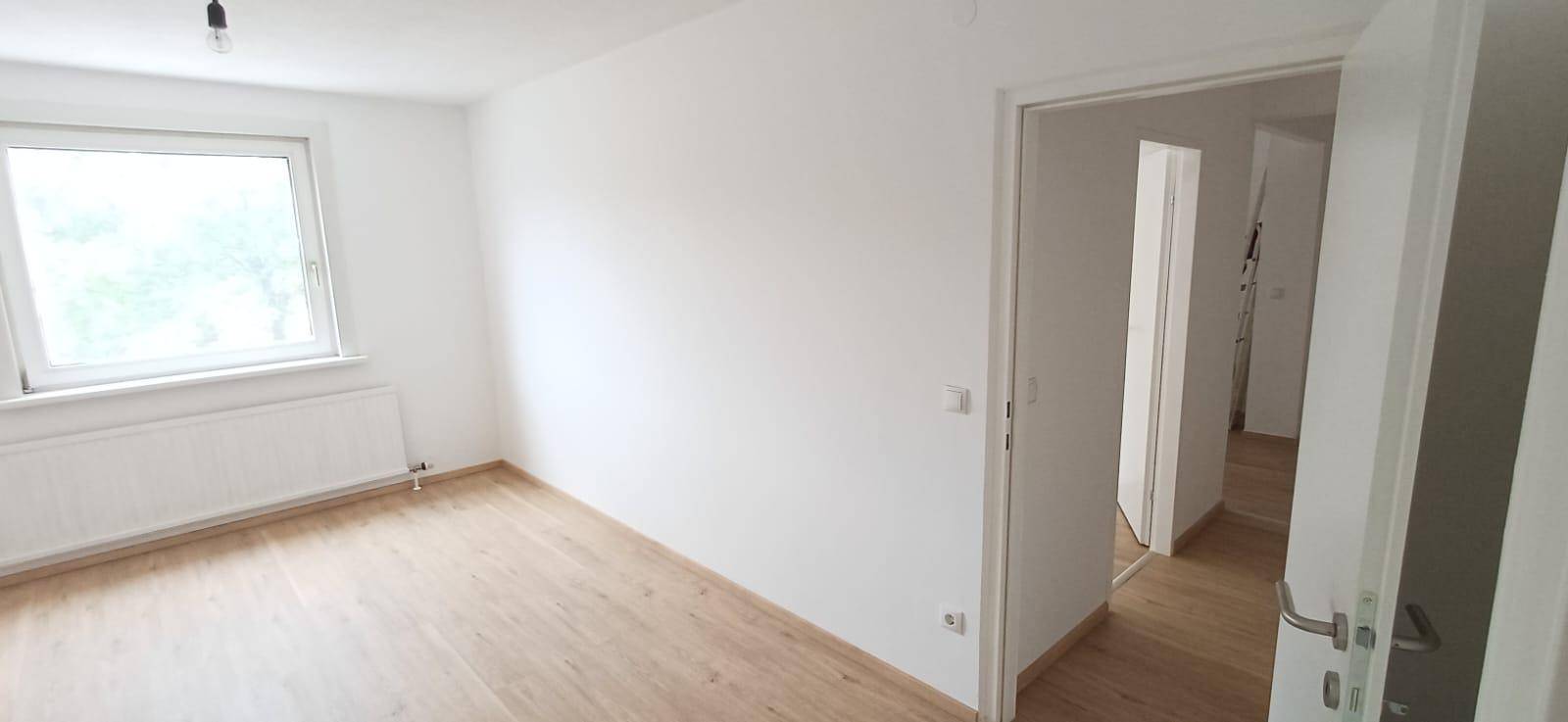 Geräumiges, helles Zimmer mit Holzfußboden und großem Fenster, das viel Tageslicht hereinlässt.