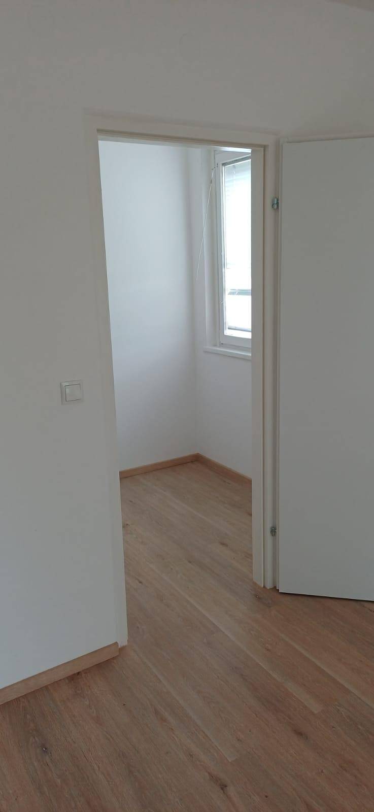 Blick durch eine offene Tür in einen weiteren hellen Raum mit Holzfußboden und Fenster.
