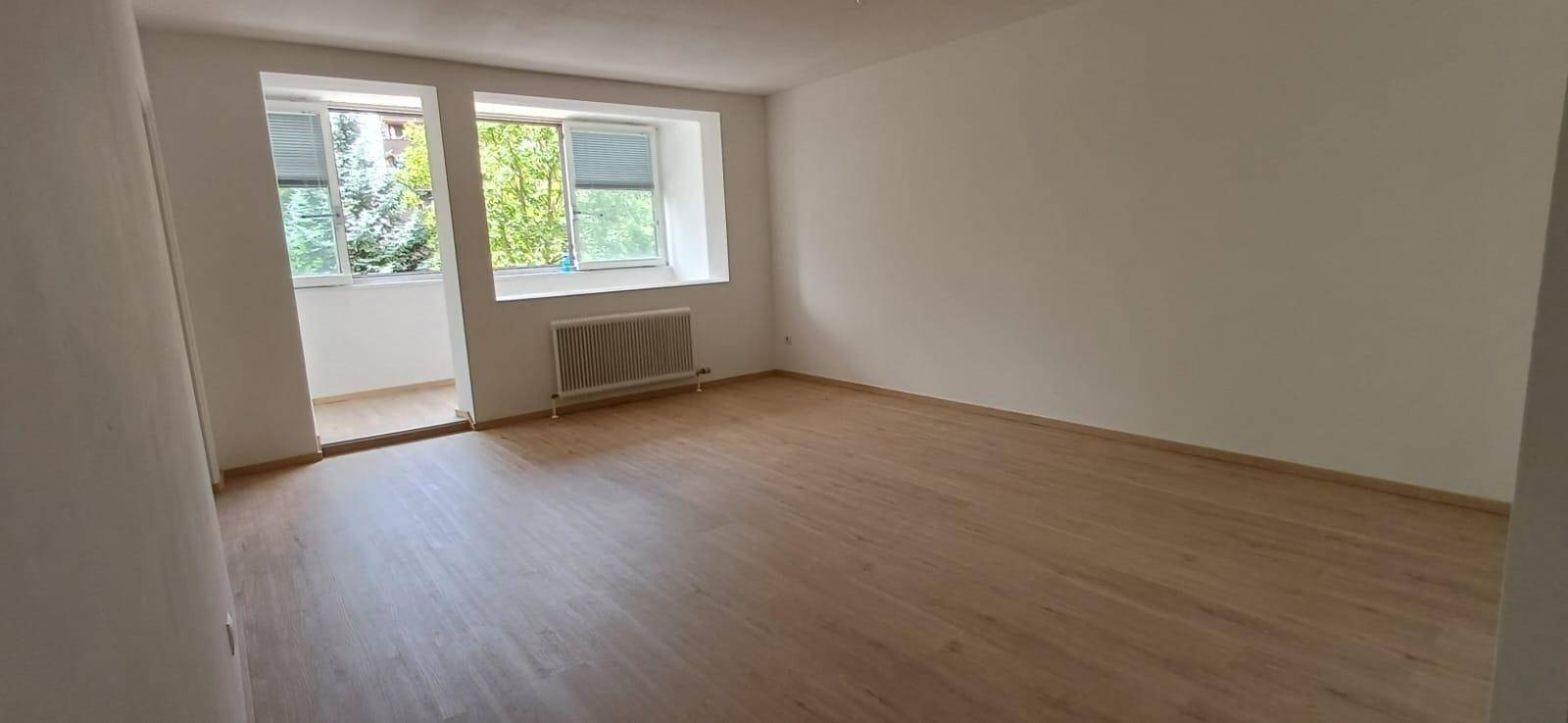 Helles Wohnzimmer mit Holzfußboden und großen Fenstern, die Zugang zu einem verglasten Balkon bieten.