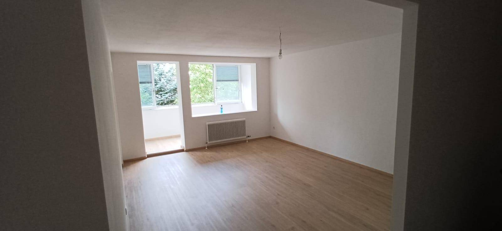Helles Wohnzimmer mit Holzfußboden und großen Fenstern, die Zugang zu einem verglasten Balkon bieten.