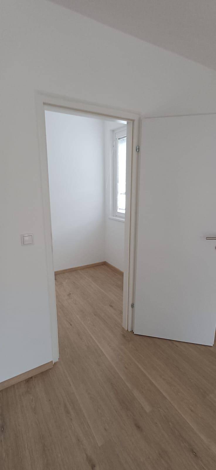 Blick durch eine offene Tür in einen weiteren hellen Raum mit Holzfußboden und Fenster.
