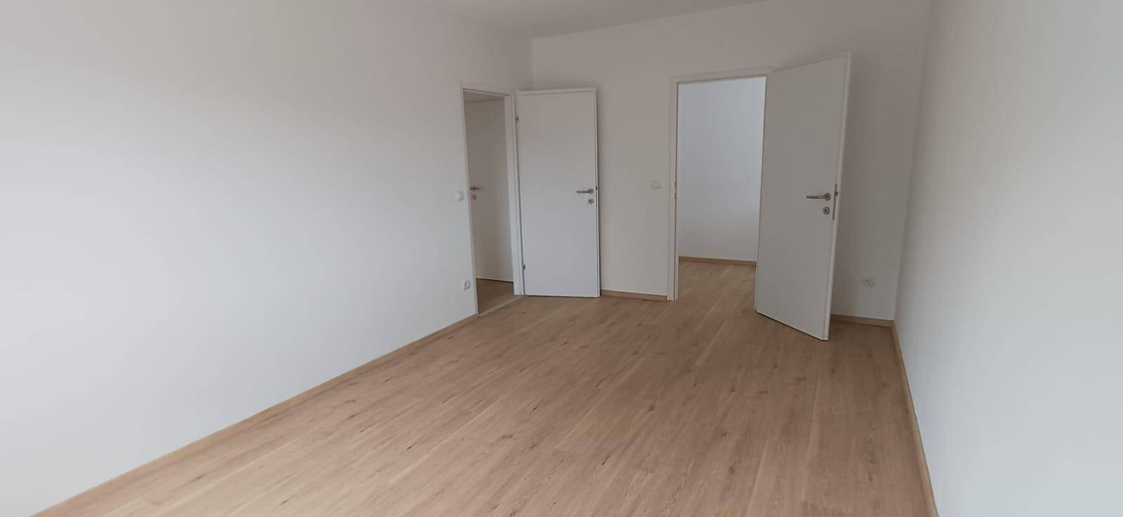 Helles Zimmer mit Holzfußboden und weißen Wänden, das Zugang zu weiteren Räumen bietet.