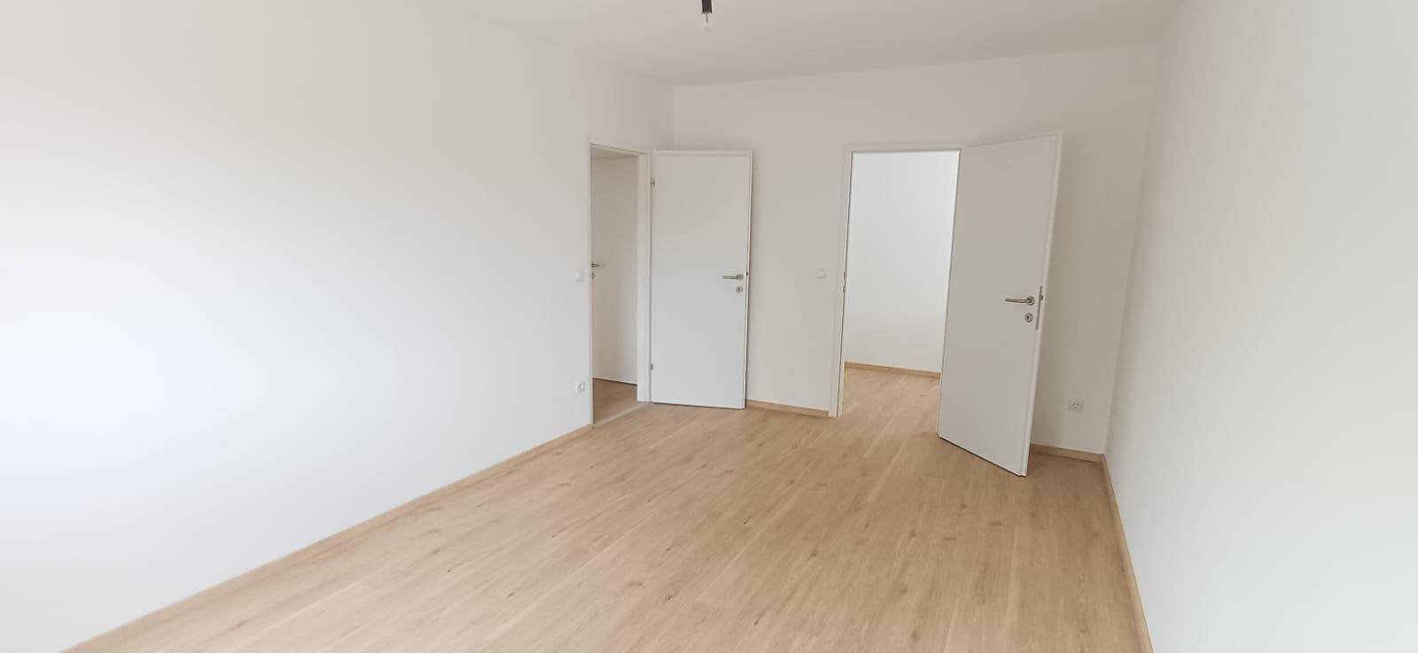Helles Zimmer mit Holzfußboden und weißen Wänden, das Zugang zu weiteren Räumen bietet.