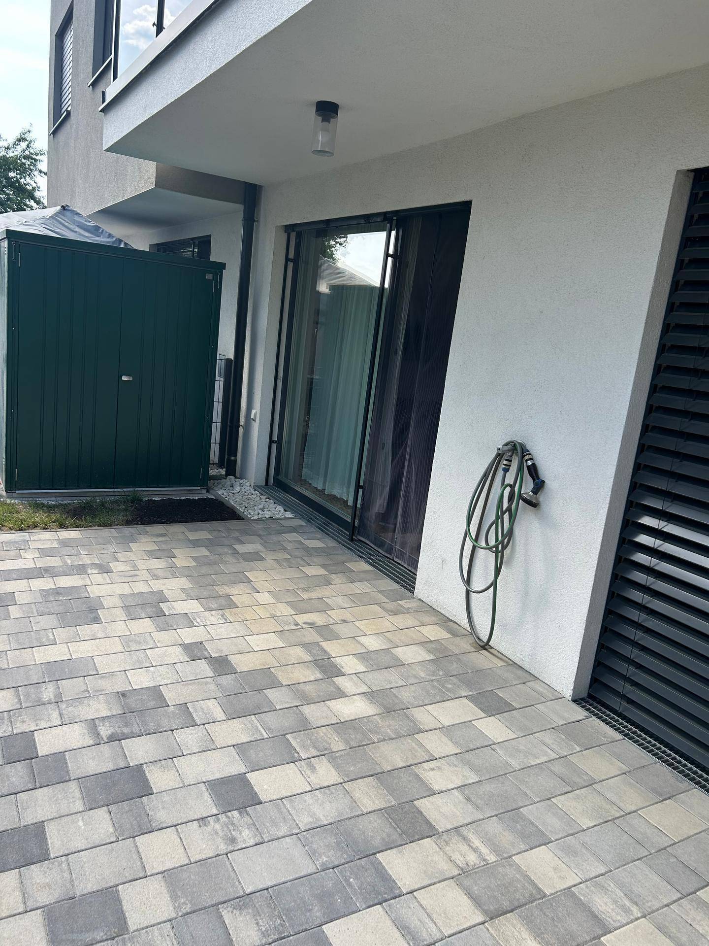 Geräumige Terrasse mit Pflastersteinen neben dem Gebäude, im Hintergrund ein grüner Gartenschuppen.