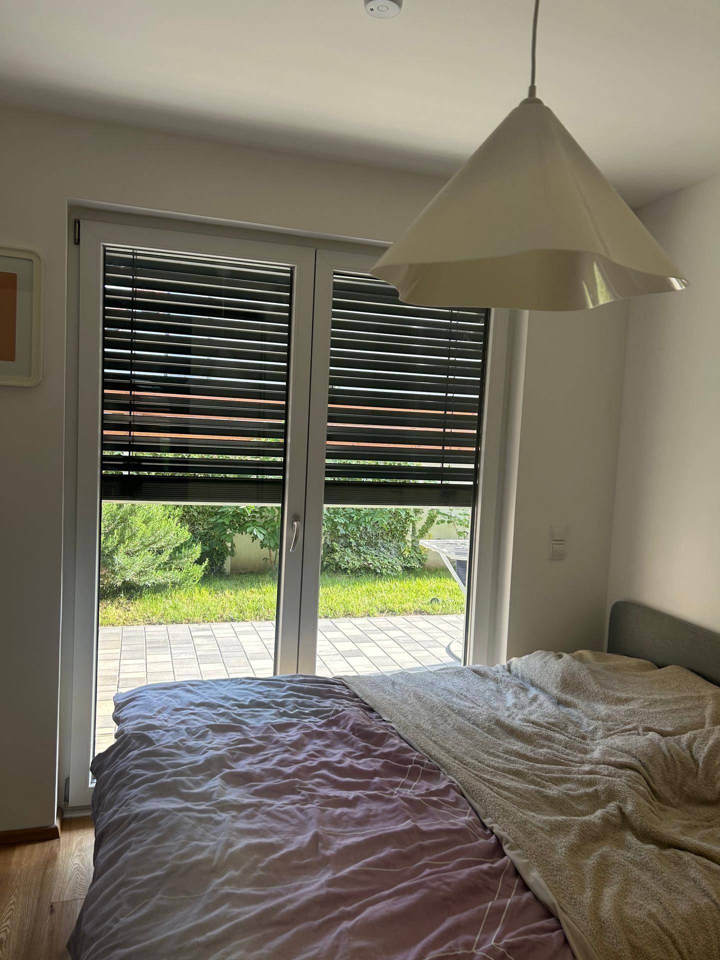 Schlafzimmerfenster mit geschlossenen Jalousien, das einen Blick auf den grünen Garten freigibt.