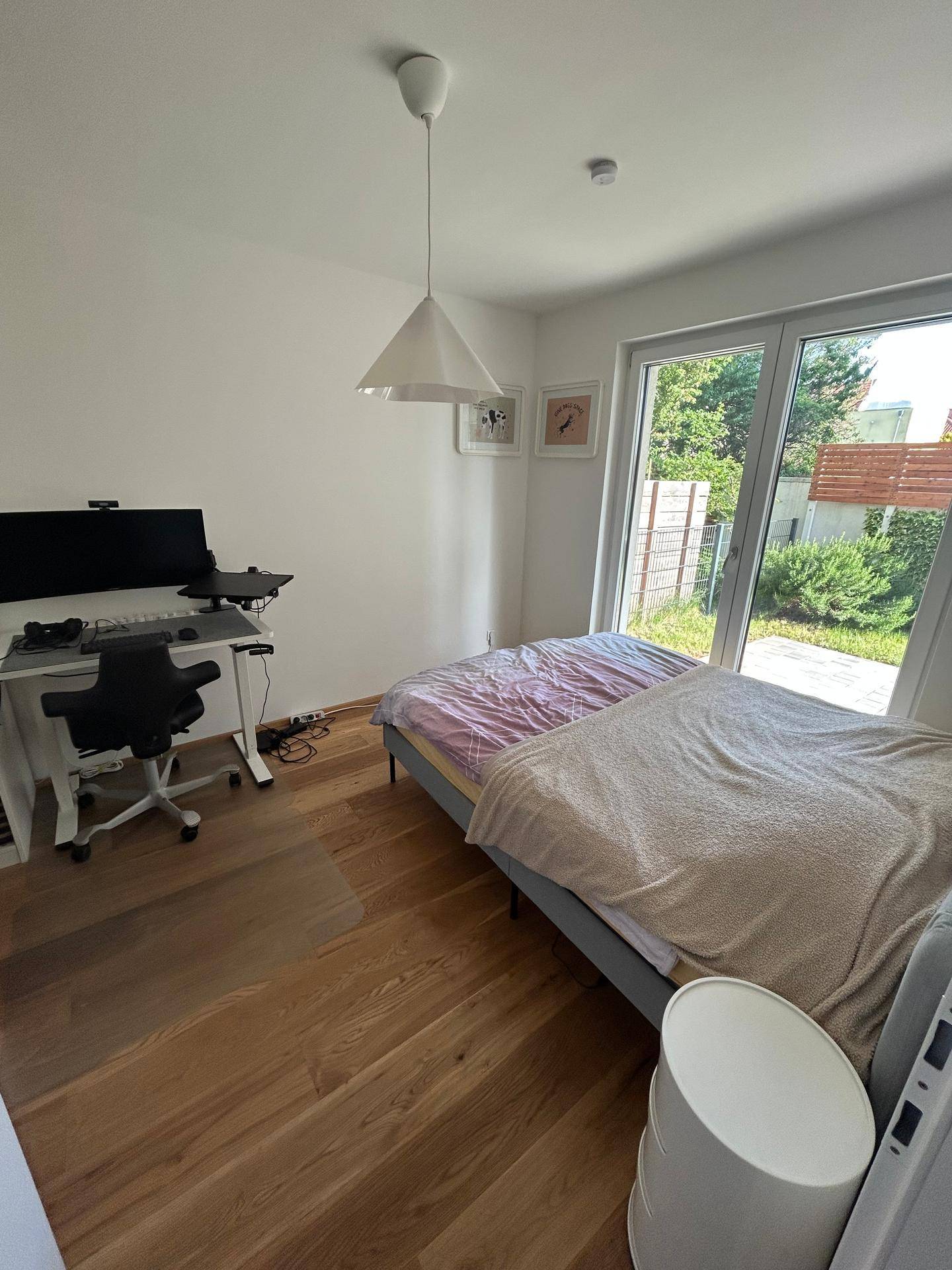 Helles Schlafzimmer mit Holzboden, Schreibtisch und großem Fenster mit Zugang zum Außenbereich.