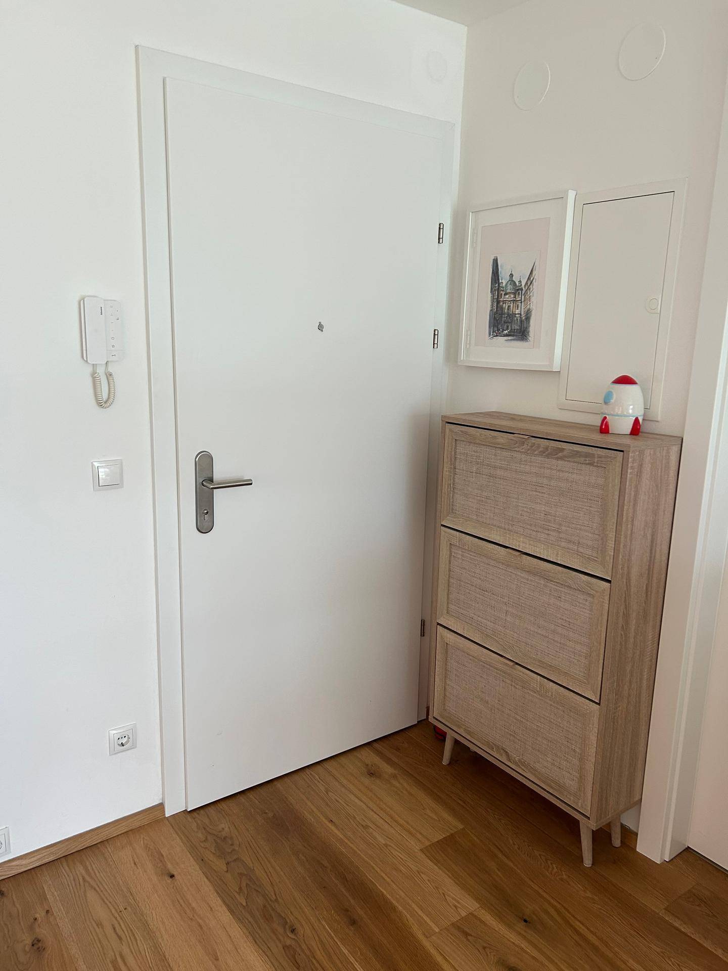 Heller Eingangsbereich mit Holzboden, weißer Tür und einem kleinen, modernen Schuhschrank.