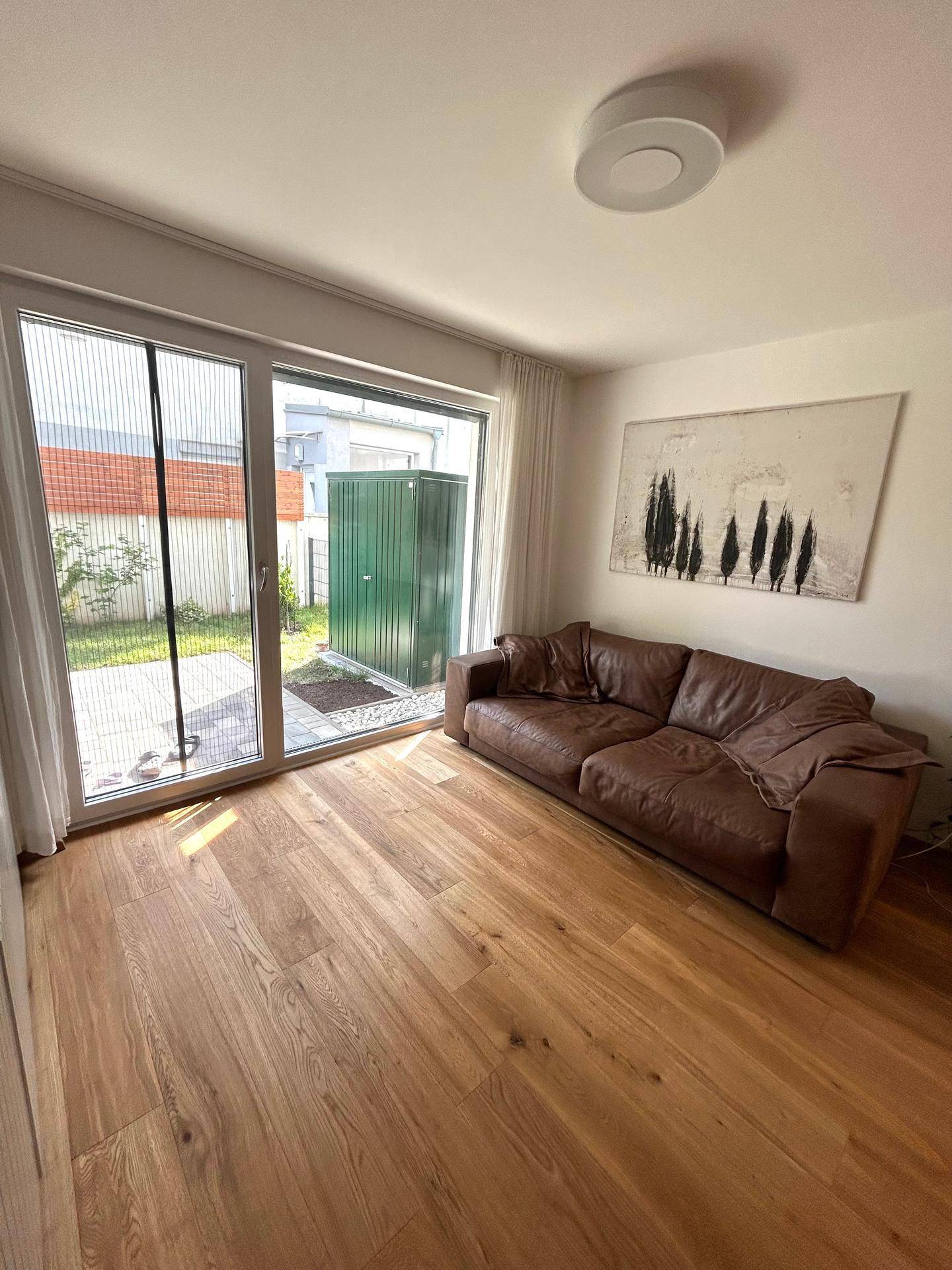 Helles Wohnzimmer mit braunem Ledersofa, Holzboden und Zugang zur Terrasse durch eine große Schiebetür.