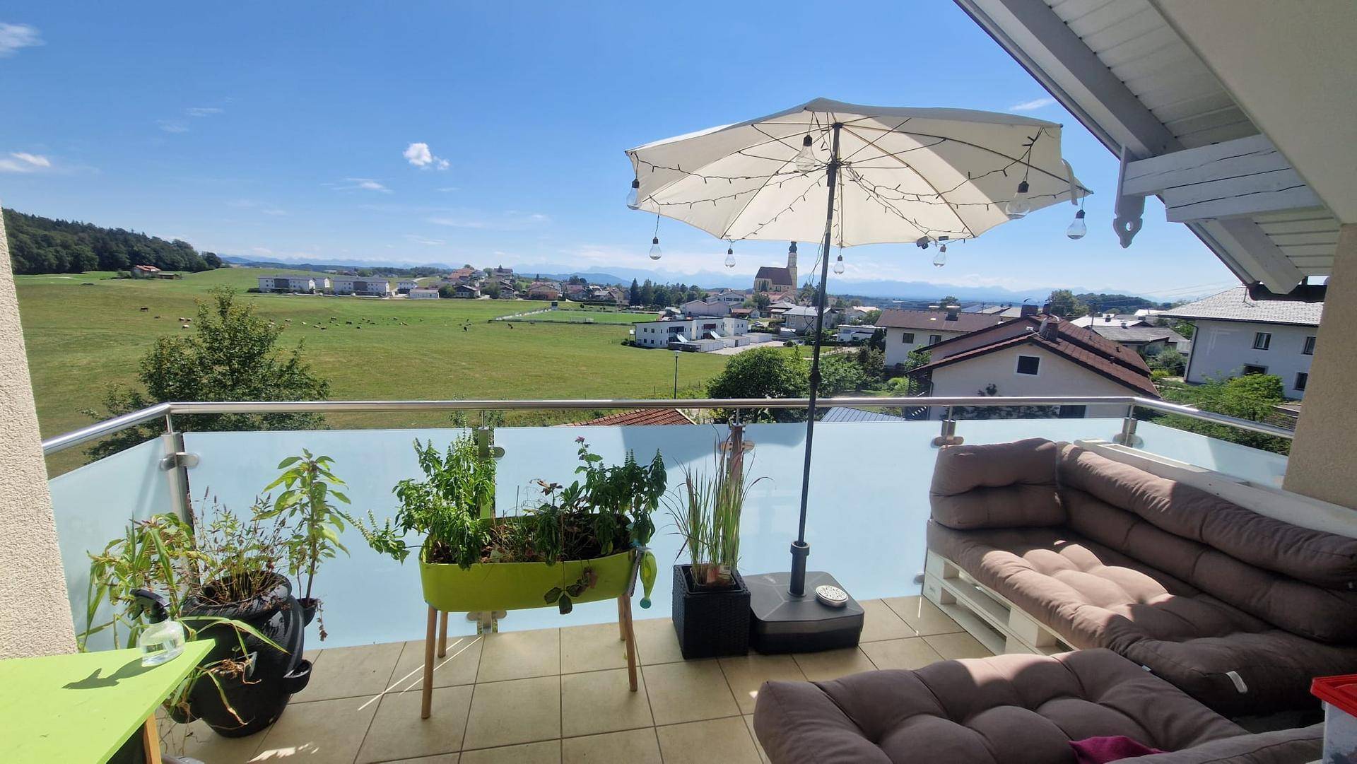 Heller Balkon mit Sitzgelegenheiten, Pflanzen und Panoramablick über Felder und Berge.