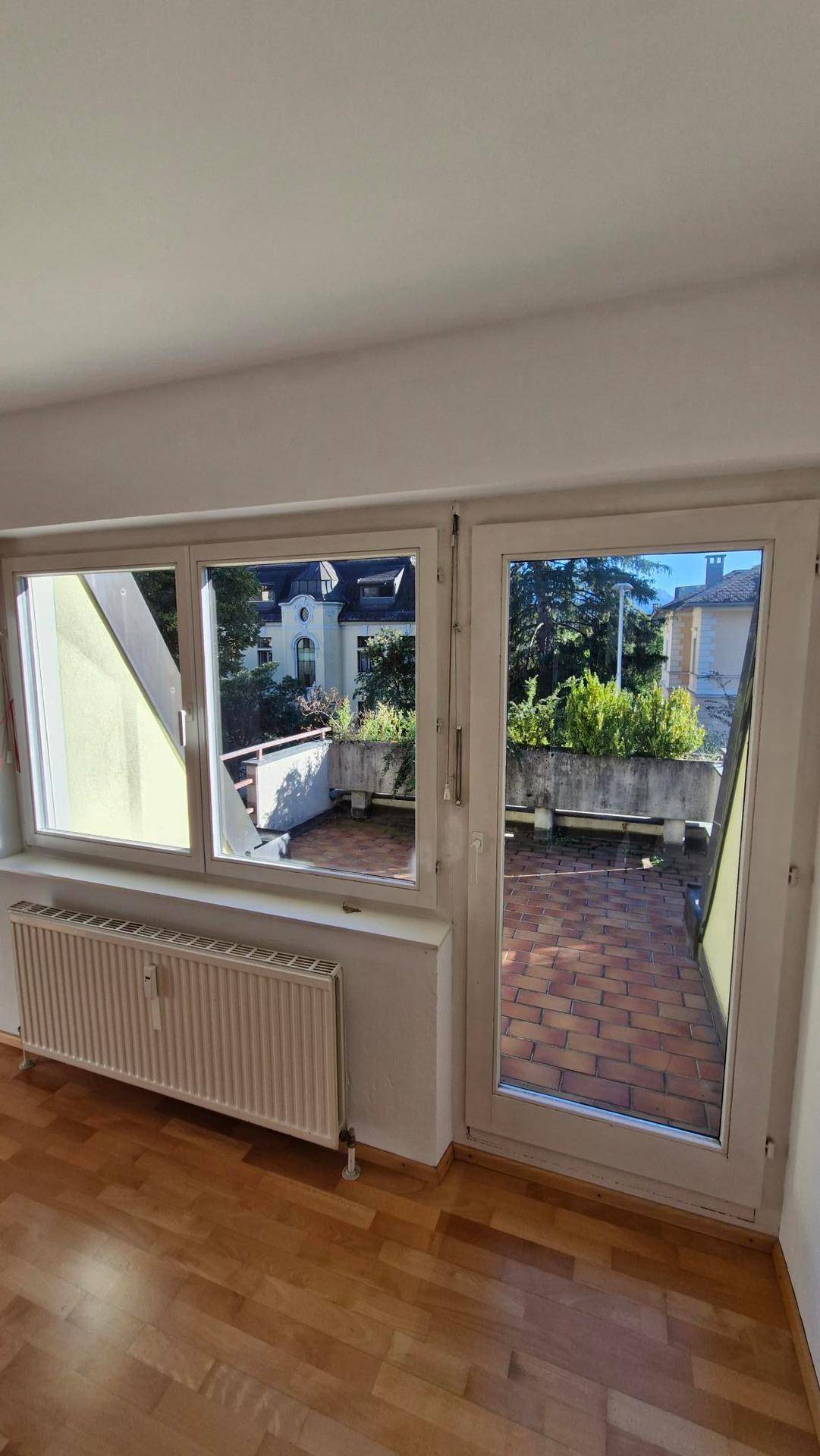 Blick auf eine geflieste Terrasse mit Pflanzen und umliegenden Gebäuden von einem hellen Zimmer aus.