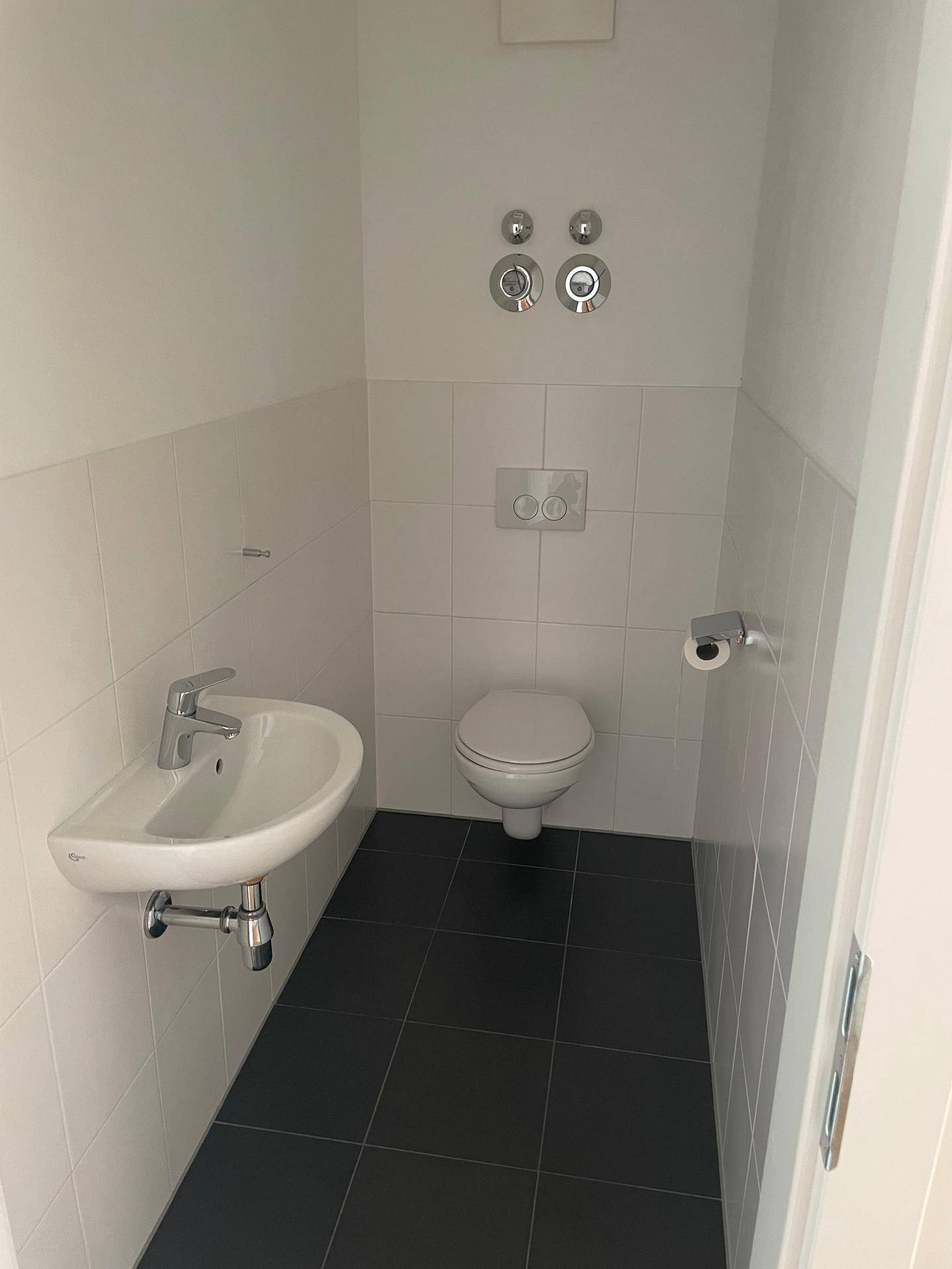 Separates Gäste-WC mit Wand-WC, kleinem Waschbecken und modernen weißen sowie dunklen Fliesen.